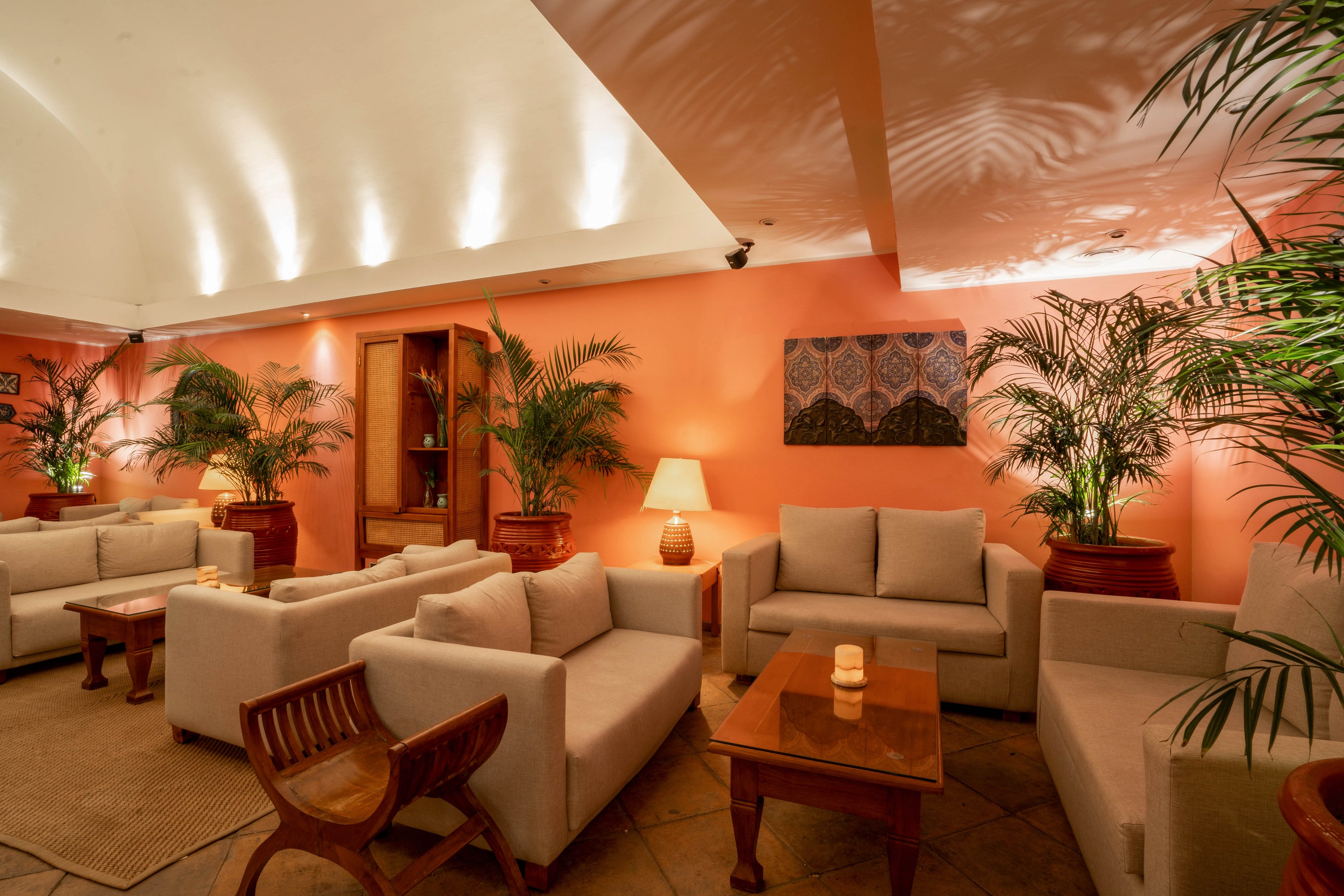 A warmly lit lounge features modern beige sofas arranged around wooden tables, creating an inviting seating area. The walls are painted a soft peach, complemented by large potted plants and ambient table lamps. A piece of abstract wall art adds visual interest, while candles on the tables enhance the cozy atmosphere. The setting suggests a relaxed, comfortable environment ideal for socializing or unwinding.