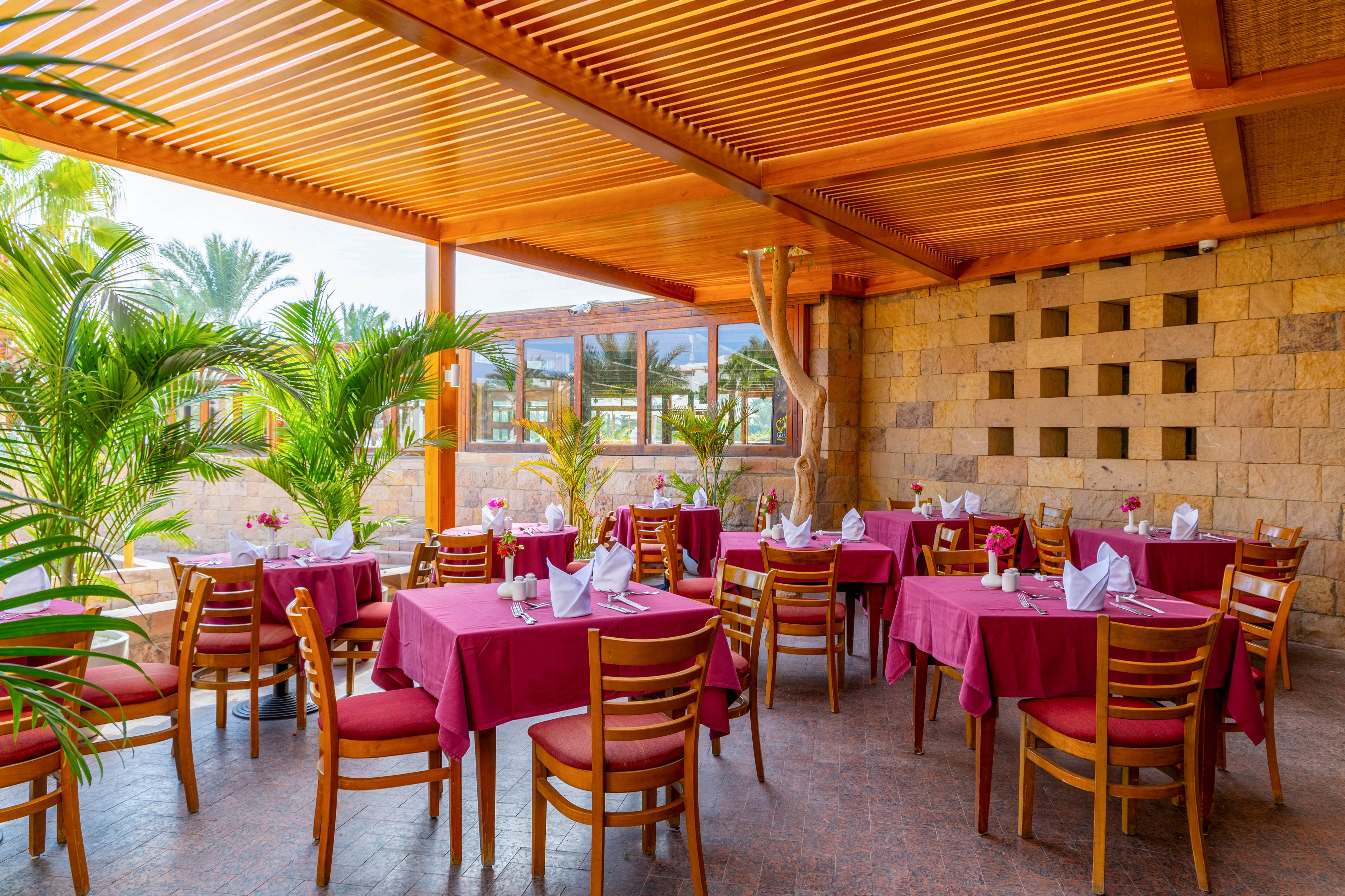 An open-air restaurant patio features wooden tables covered with red tablecloths and neatly folded napkins. Wooden chairs are arranged around each table, and lush green plants add a tropical touch to the setting. The space is shaded by a wooden slatted ceiling, with natural light streaming in. The background includes stone walls and large windows, creating a relaxed and inviting atmosphere. No visible text or numbers are present in the image.