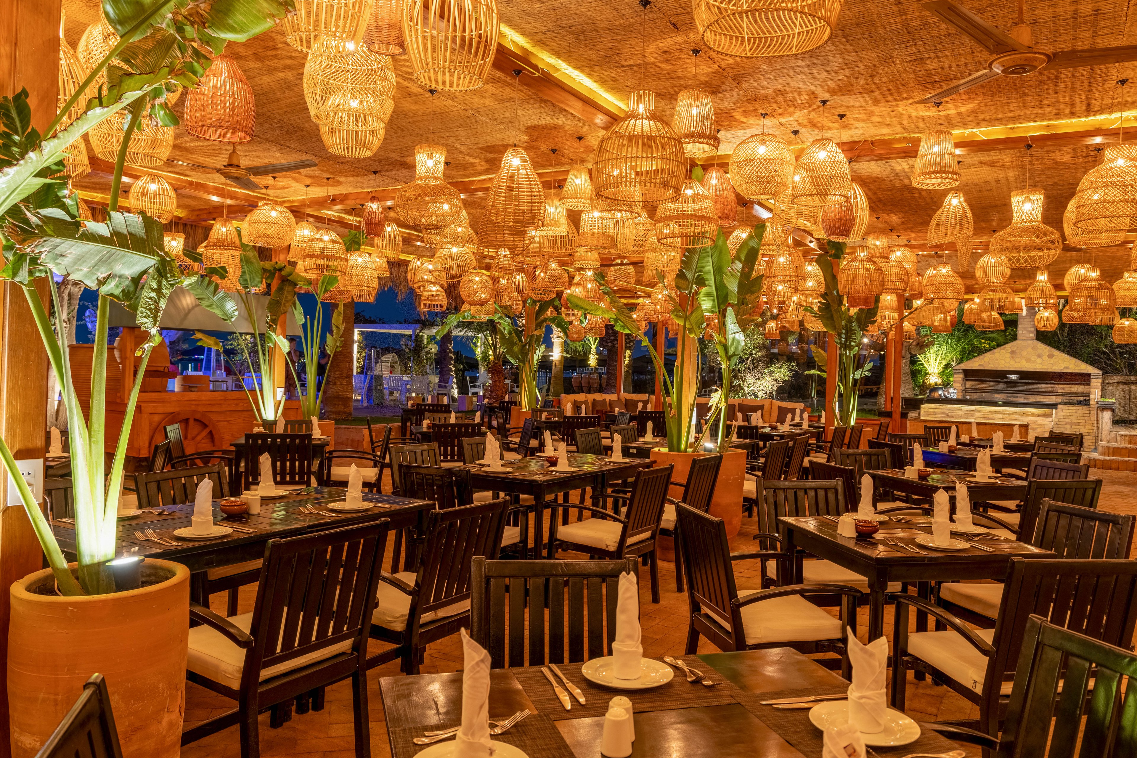 A spacious restaurant dining area features dark wooden tables and chairs, each set with neatly folded napkins and tableware. The ceiling is adorned with numerous woven pendant lights, casting a warm golden glow throughout the space. Large potted plants add a lush, tropical ambiance, while the overall decor blends natural materials with a cozy, inviting atmosphere. No visible people or text are present in the image.