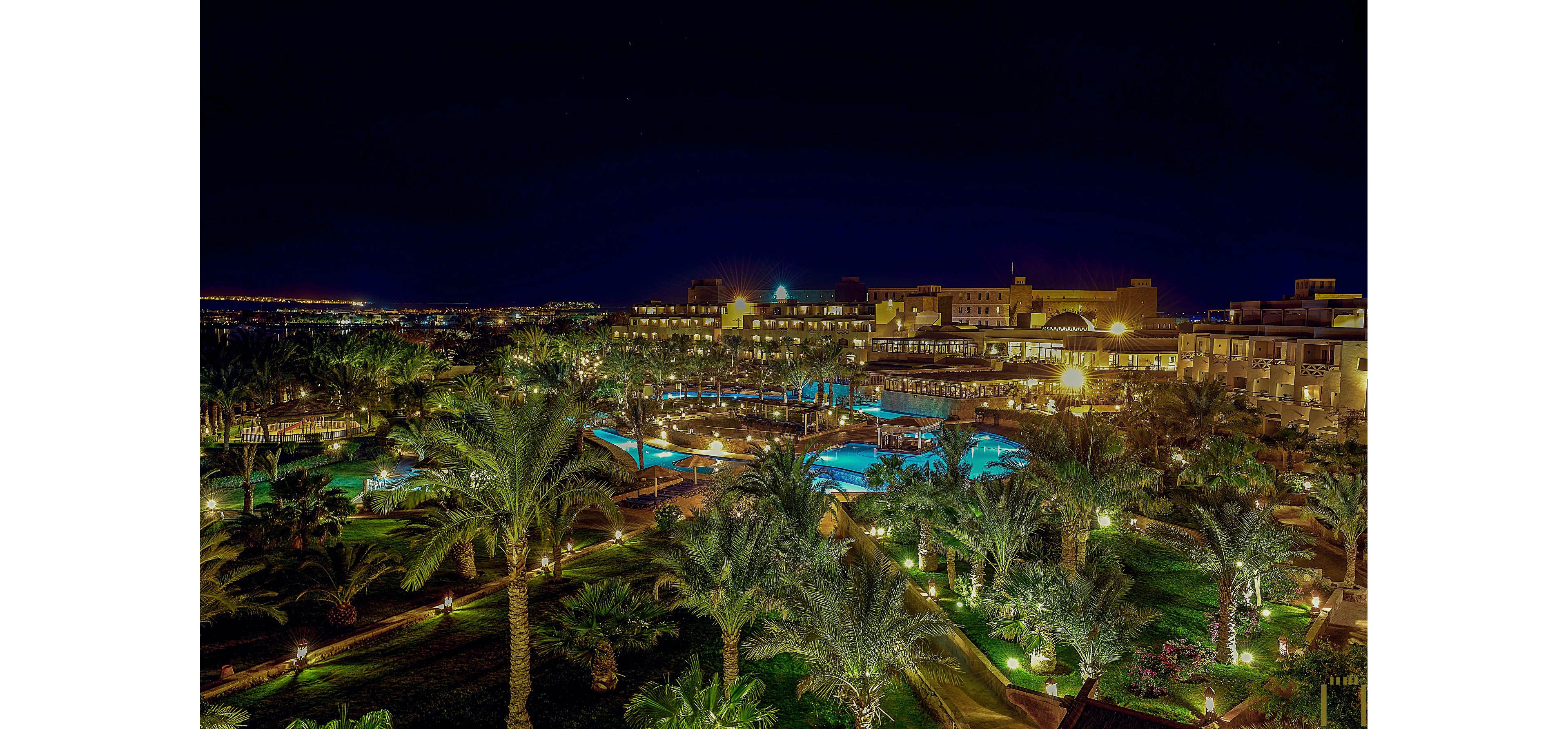 A sprawling luxury resort is beautifully lit up at night, featuring multiple swimming pools surrounded by lush palm trees and manicured gardens. The scene captures the tranquil ambiance of the resort grounds, with glowing pathways and reflections in the water. The dark sky contrasts with the vibrant, warm lighting, creating an inviting and serene atmosphere. No visible text or numeric values are present in the image.