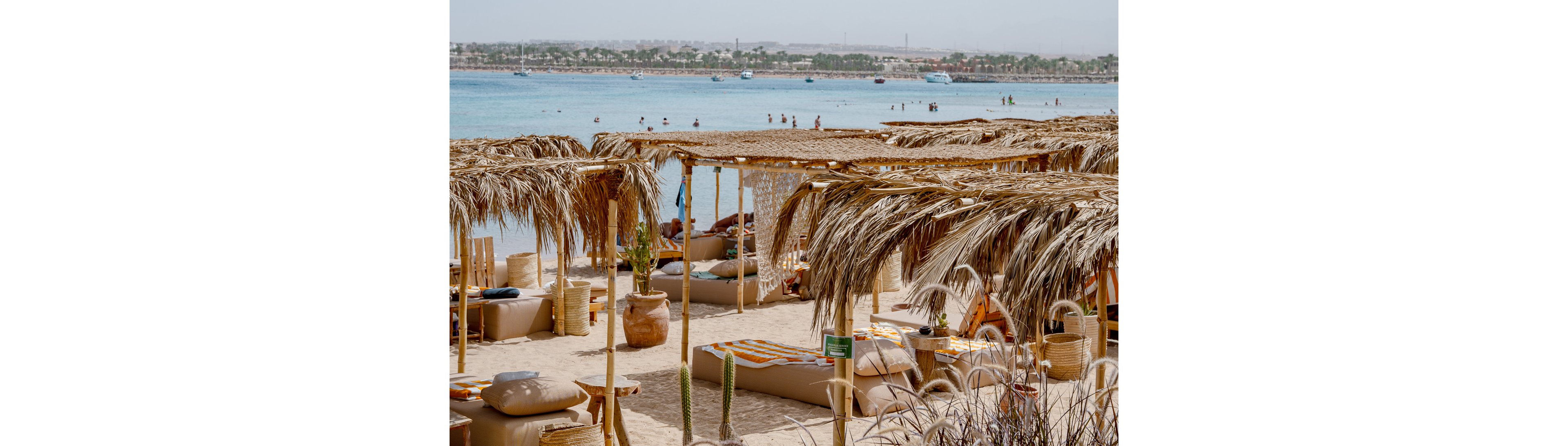 A tranquil beach scene features straw cabanas and cushioned lounge beds arranged on soft sand. The setting overlooks a calm blue sea with people swimming and relaxing in the shallow water. The atmosphere is relaxed and summery, with natural tones and clear skies. Decorative vases and grasses add to the laid-back, tropical vibe. No visible text or numbers are present in the image.