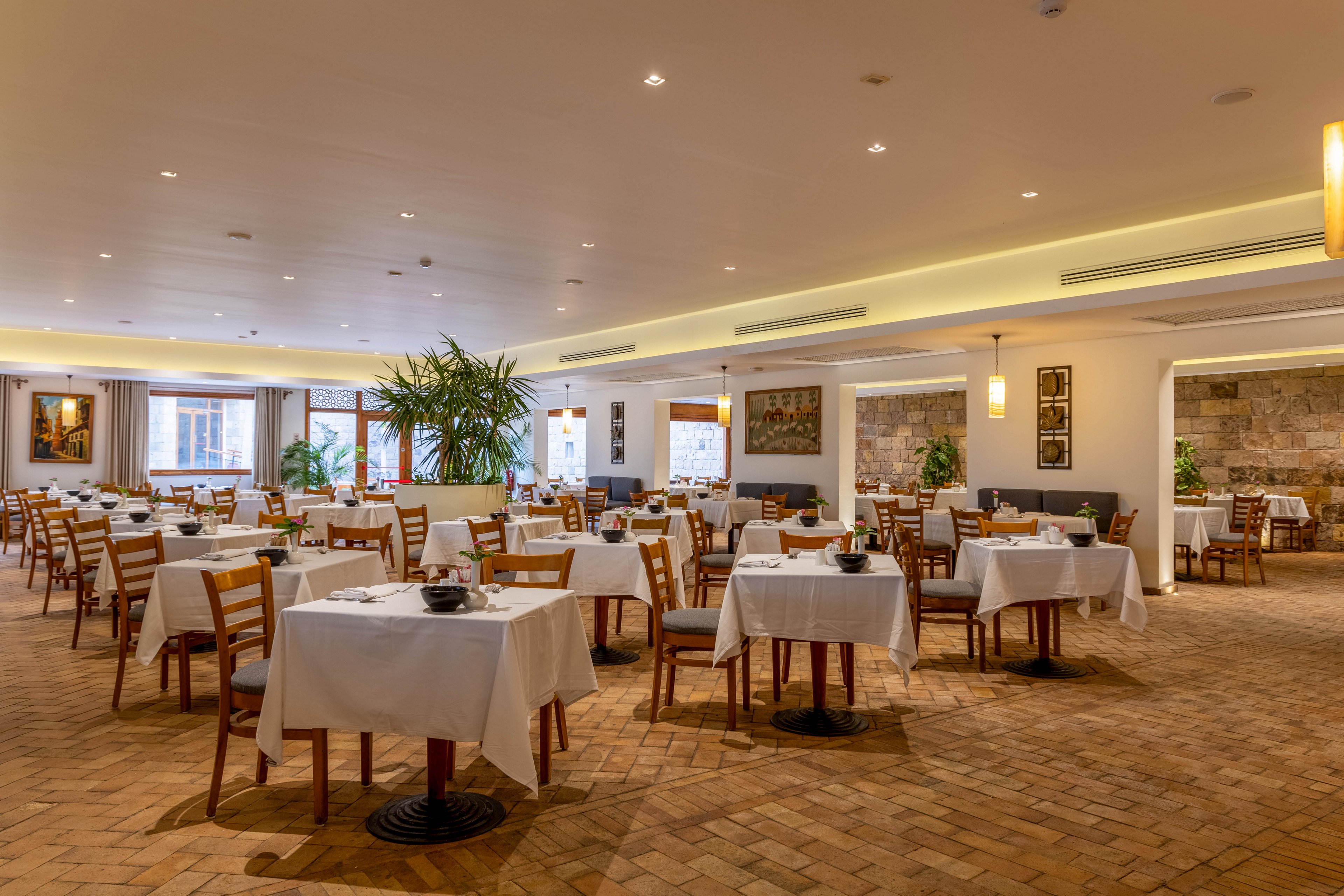 A well-lit restaurant dining area features neatly arranged tables with white tablecloths and wooden chairs. The setting is contemporary, with warm lighting, indoor plants, and artwork on the walls. The space appears clean and inviting, with a neutral color palette and a relaxed atmosphere. No people are present, emphasizing the calm and readiness for guests.
