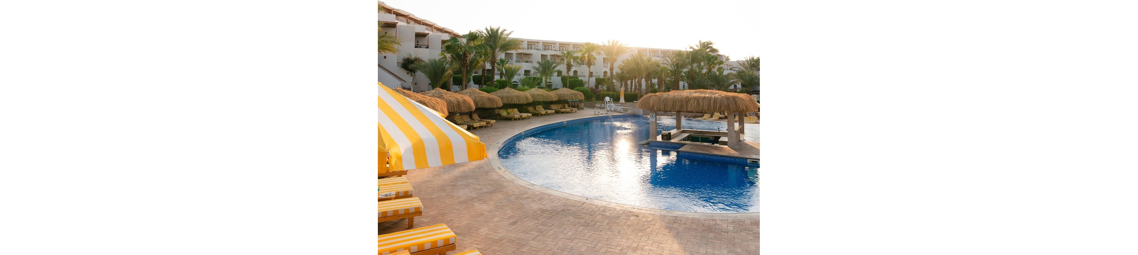 A serene resort pool area is shown at sunrise, featuring yellow and white striped loungers and umbrellas. The pool is surrounded by palm trees and thatched cabanas, with white buildings in the background. The scene is bathed in warm, golden light, creating a relaxed and inviting atmosphere. No visible people or text are present in the image.