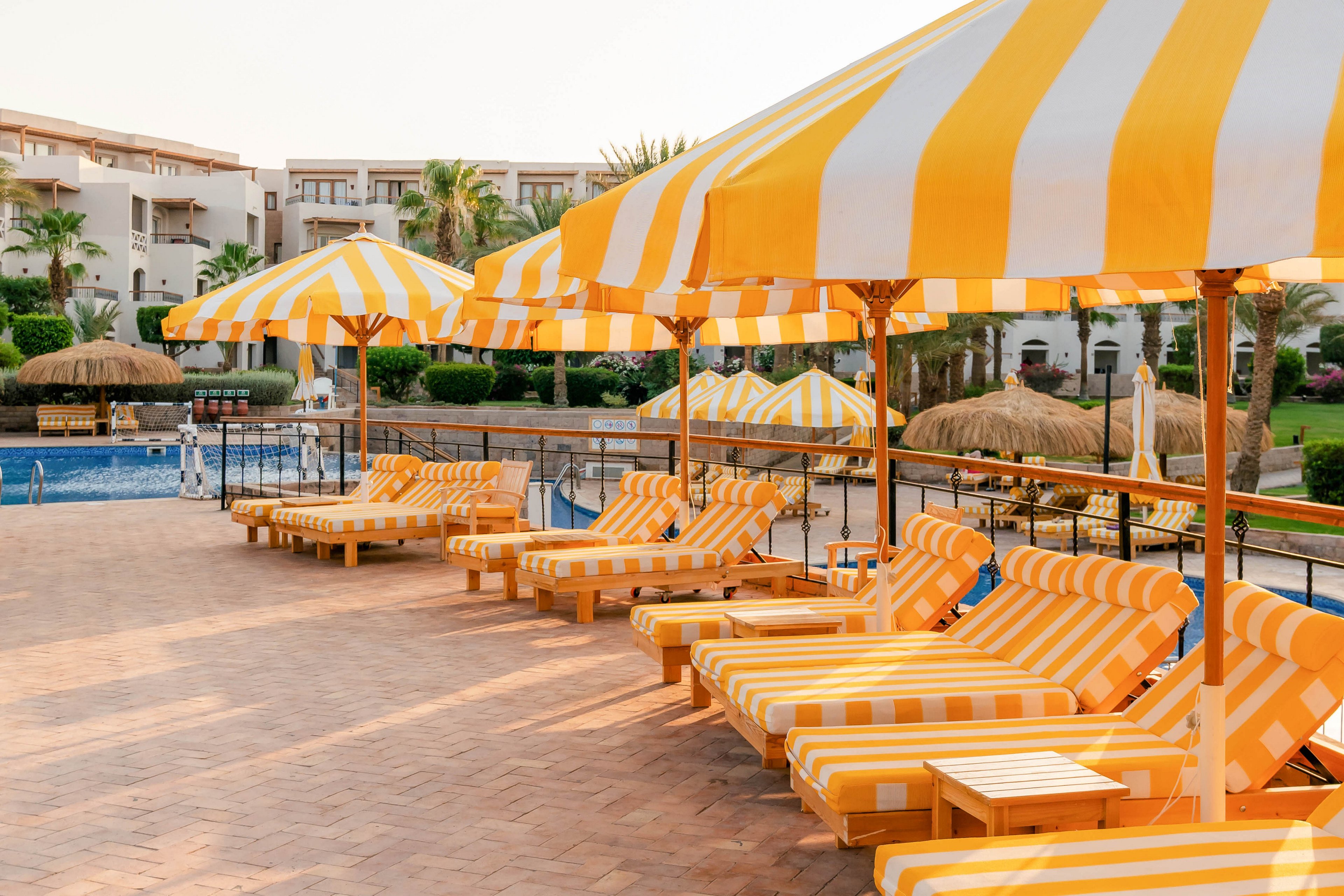 A row of yellow and white striped lounge chairs and umbrellas lines a sunlit poolside terrace. The setting is an outdoor resort area with tiled flooring and a swimming pool visible in the background. The atmosphere is relaxed and inviting, with clear skies and warm lighting. No people are present, emphasizing a tranquil vacation vibe. The image features a harmonious yellow and white color palette.