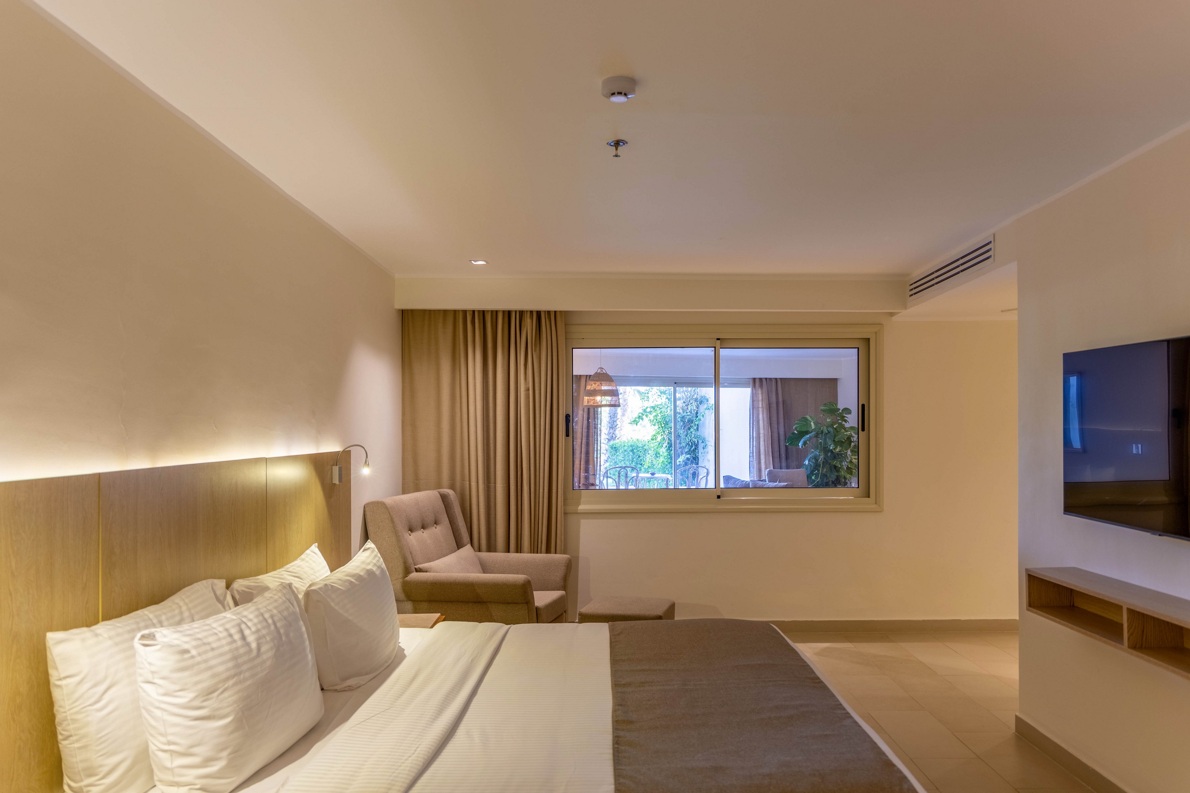 A contemporary hotel room features a neatly made bed with white linens, a cushioned armchair, and a wall-mounted television. The space is illuminated by soft, warm lighting and natural light from a large window overlooking greenery. Neutral tones and minimalist decor create a calm, inviting atmosphere.