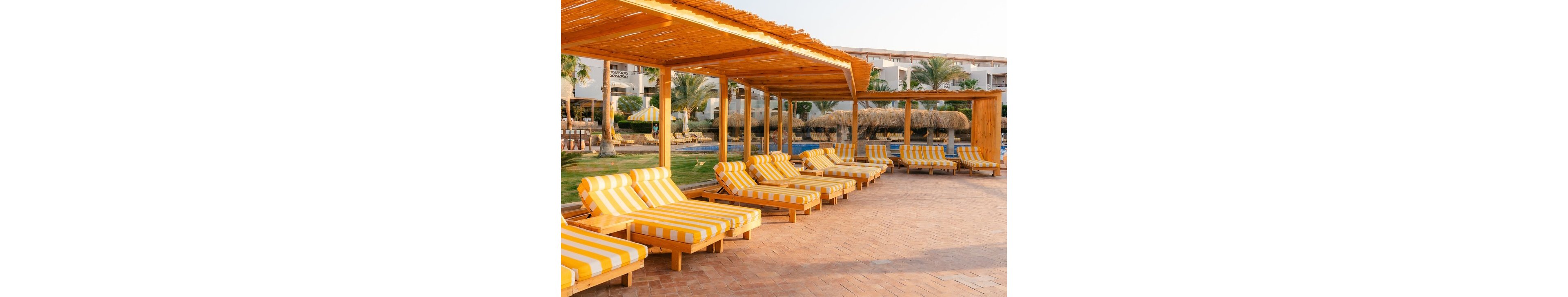 A row of yellow and white striped lounge chairs is arranged under a wooden pergola beside a resort pool. The setting is outdoors with palm trees, manicured lawns, and a clear sky, evoking a relaxed vacation atmosphere. The area is paved with terracotta tiles, and the resort buildings are visible in the background. The overall mood is bright, inviting, and tranquil.