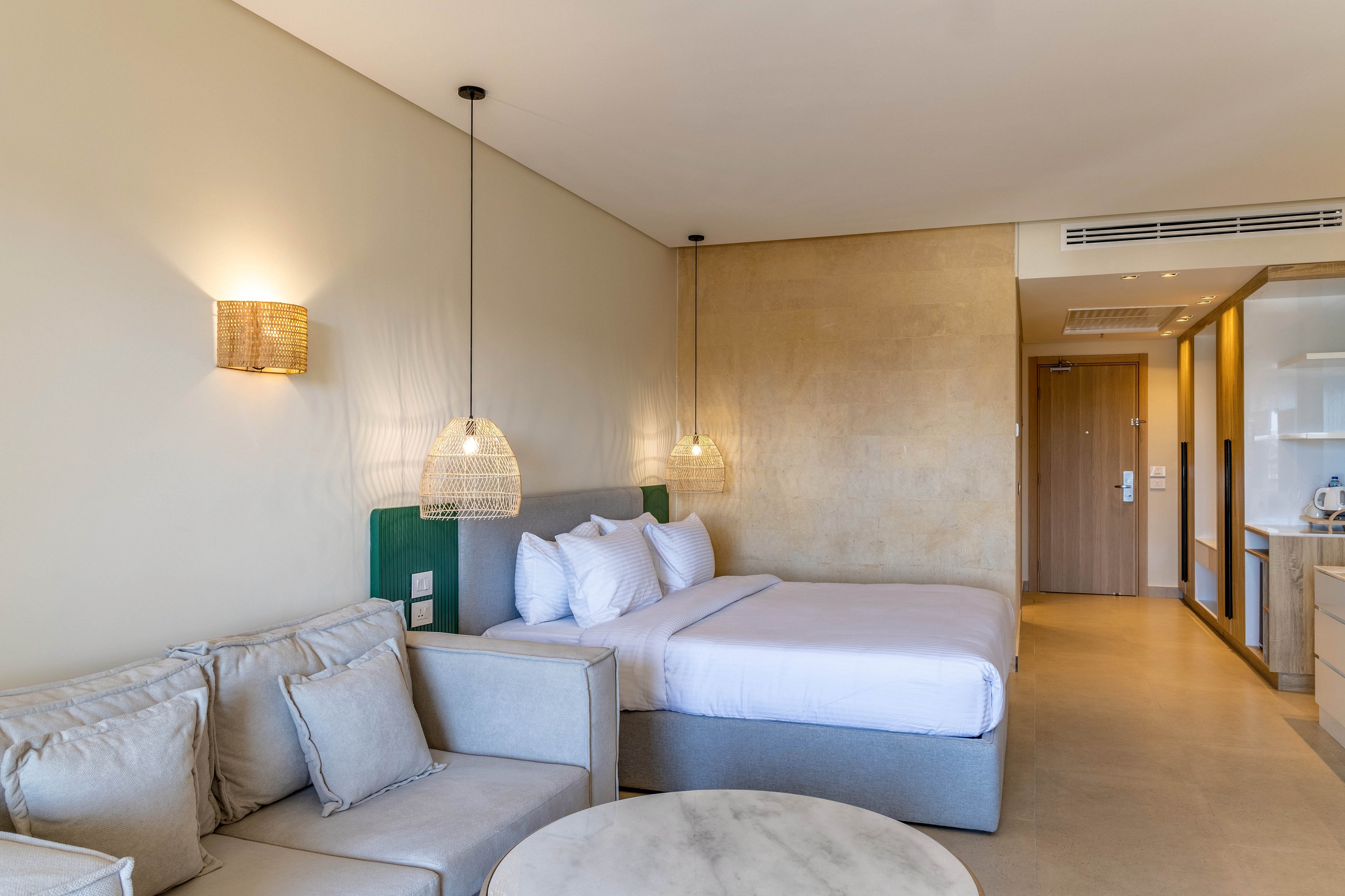 A spacious hotel room features a large bed with crisp white linens and multiple pillows, complemented by a comfortable beige sofa. The setting is bright and minimalistic, with pendant lights and a neutral color palette creating a relaxing atmosphere. The room includes a small round table, built-in cabinetry, and a visible entryway. The overall style is contemporary and inviting, ideal for leisure or business travelers.