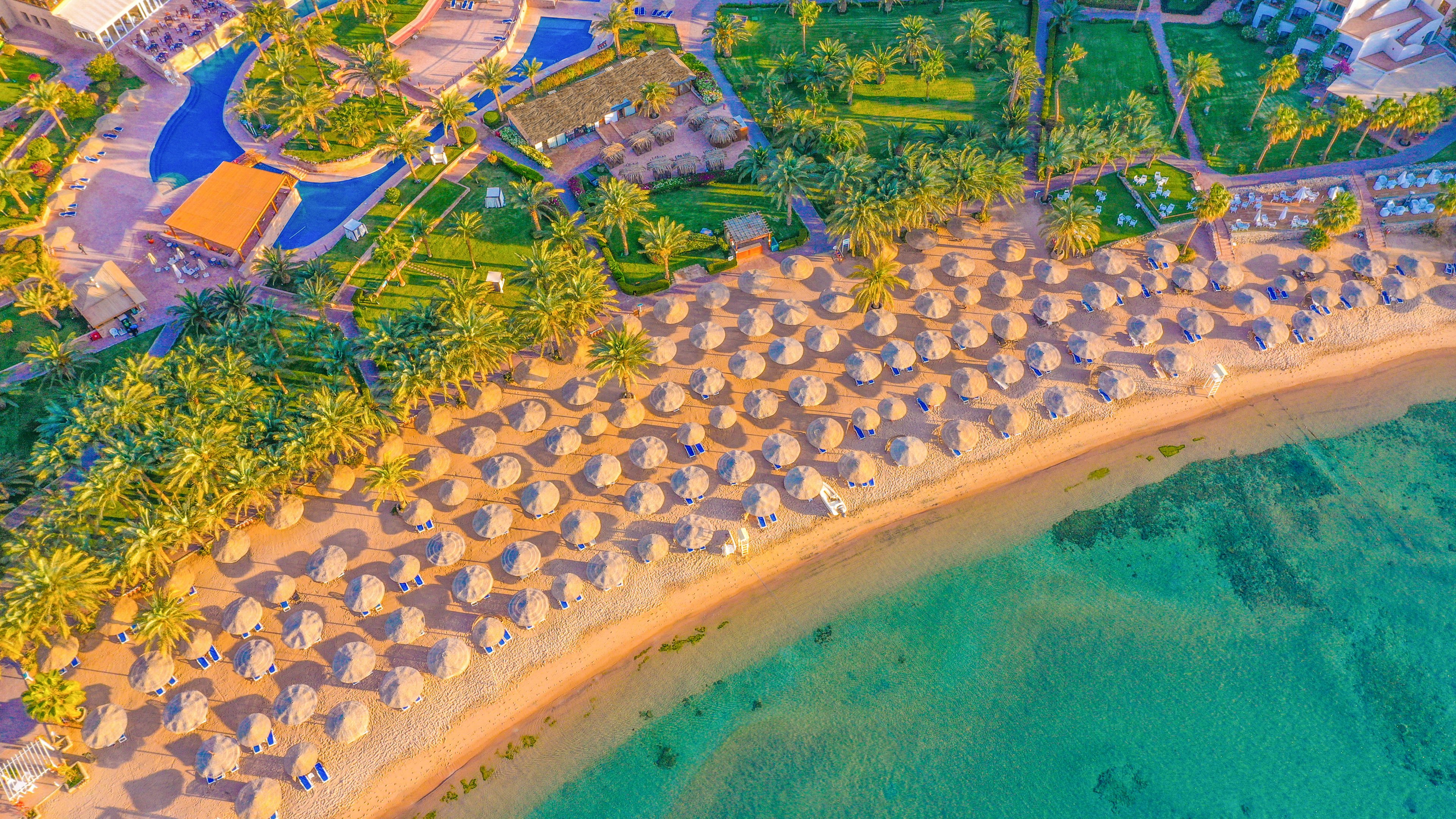 This image shows a vibrant aerial view of a tropical beach resort. Numerous straw umbrellas are neatly arranged along the sandy shoreline, with clear turquoise water gently meeting the beach. Lush palm trees and landscaped gardens surround the resort buildings, creating a relaxing and inviting atmosphere. The scene is bright and colorful, evoking a sense of leisure and vacation.