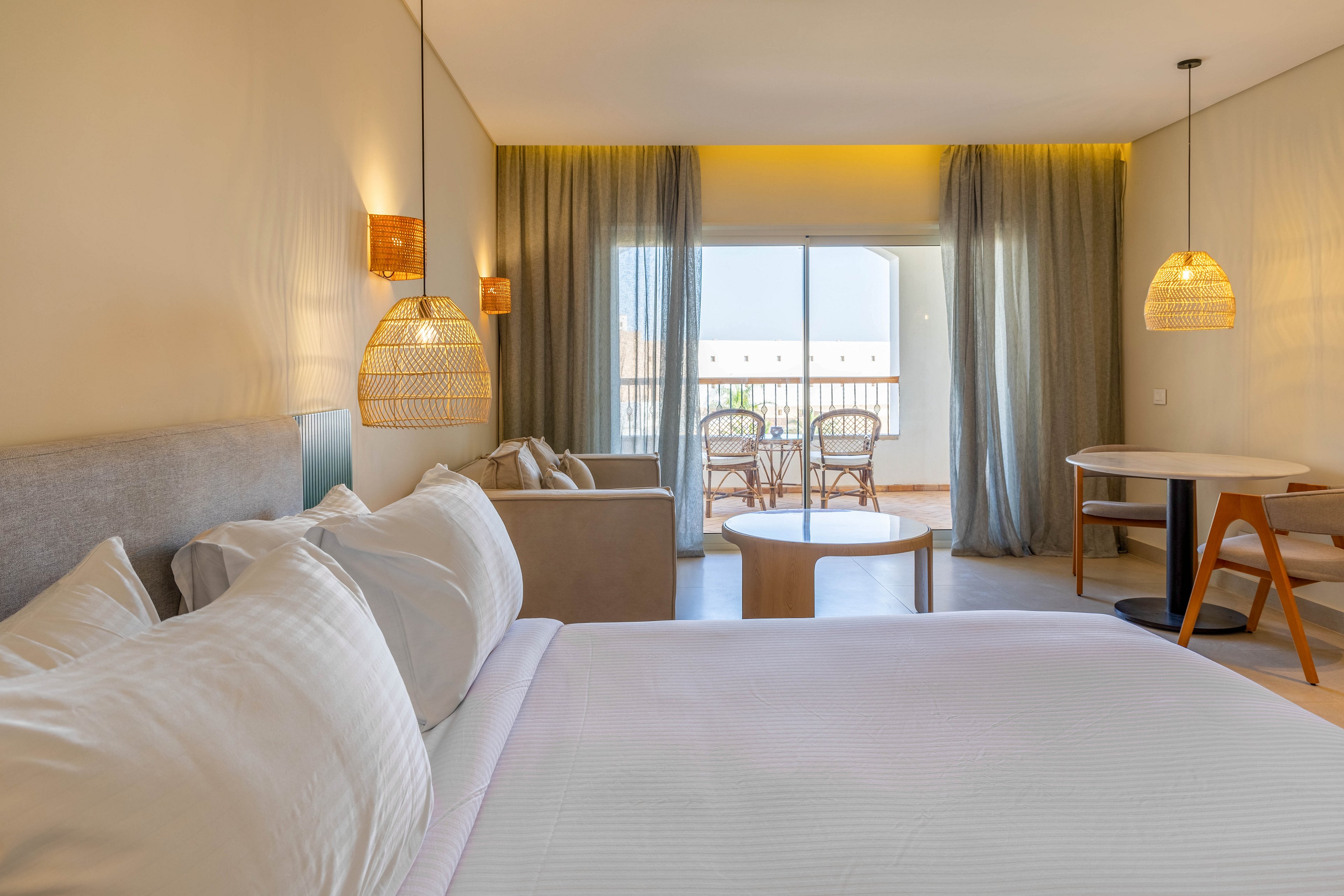 A spacious hotel room features a large bed with crisp white linens, a cozy seating area, and a round table. The room is illuminated by natural light streaming through floor-to-ceiling windows that open to a balcony with wicker chairs and a table. Warm neutral tones and woven pendant lights create a relaxing, contemporary atmosphere.