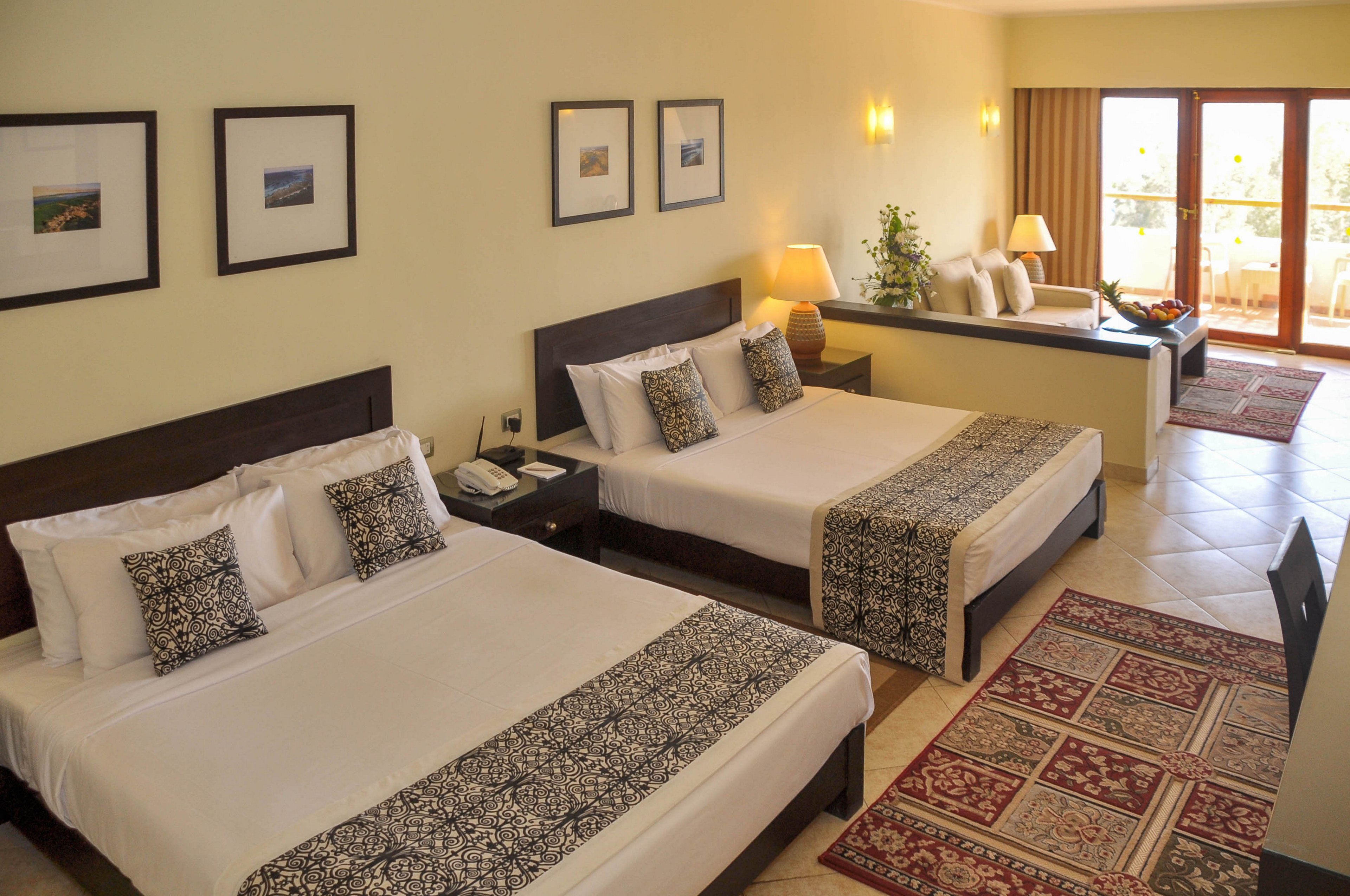 The image shows a spacious hotel room featuring two double beds with patterned runners and matching pillows. The setting includes a sitting area, desk, lamps, framed wall art, and a balcony with glass doors letting in natural light. The decor is contemporary with neutral tones and warm lighting, creating a welcoming and comfortable atmosphere. No visible text or numeric values are present in the image.