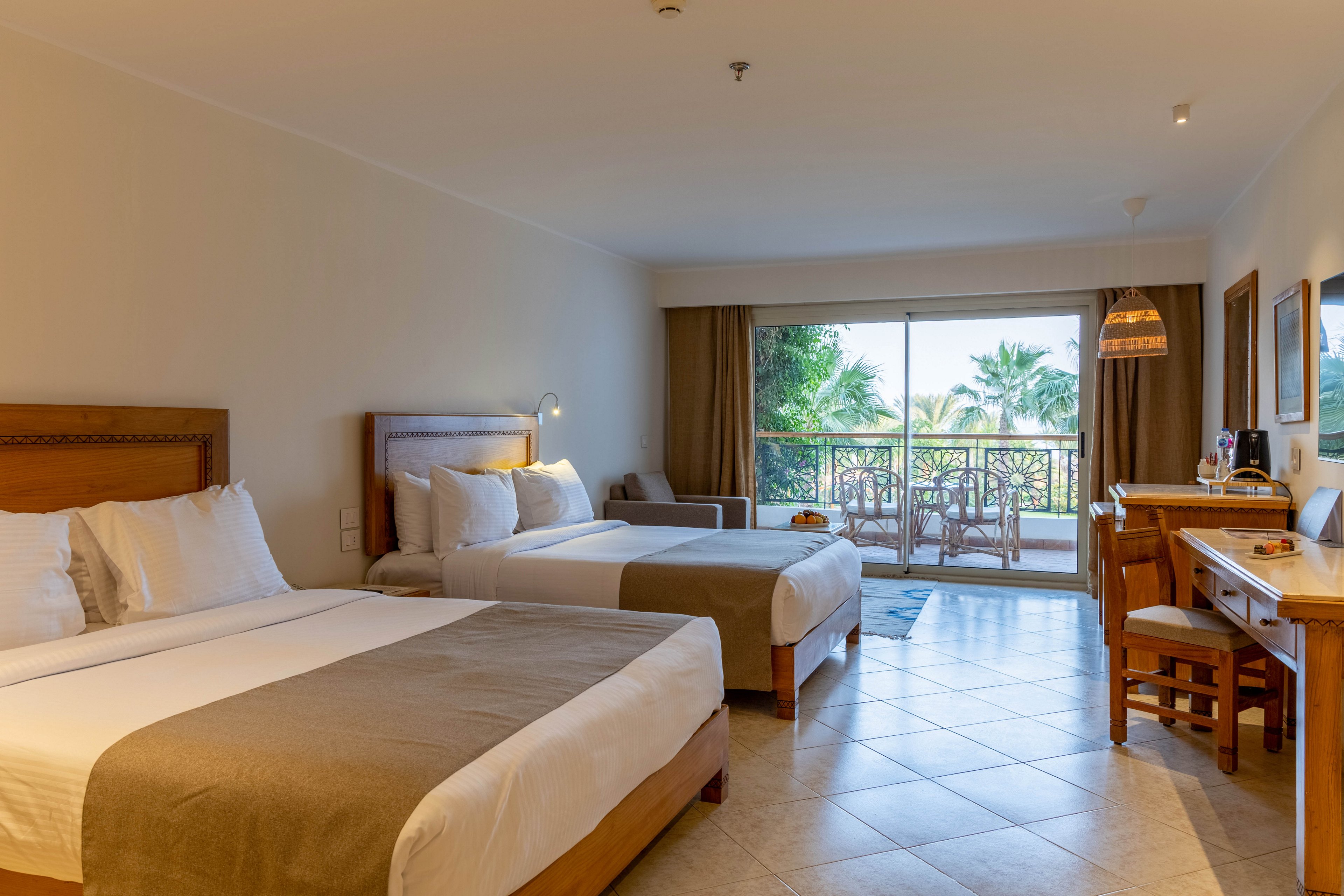A modern hotel room features two neatly made double beds with neutral bedding. The space includes a wooden desk, chairs, and a large sliding glass door leading to a balcony overlooking lush greenery. Warm lighting and tiled floors create a welcoming, relaxed atmosphere, ideal for leisure or business travelers. No visible text or numbers are present in the image.