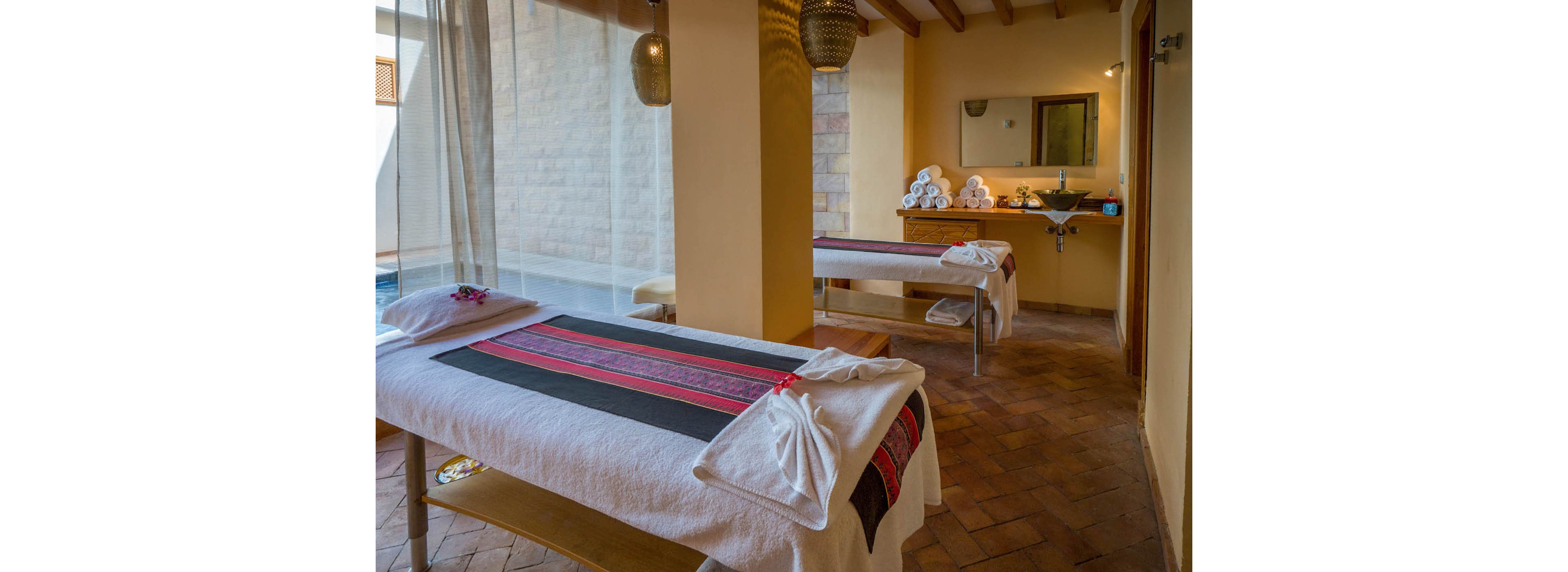 A tranquil spa room features two massage tables adorned with white linens and decorative runners. The setting is softly lit with warm tones, woven pendant lights, and a stack of rolled towels near a sink and mirror. The space exudes relaxation and comfort, with natural light filtering through sheer curtains and a peaceful, inviting atmosphere.