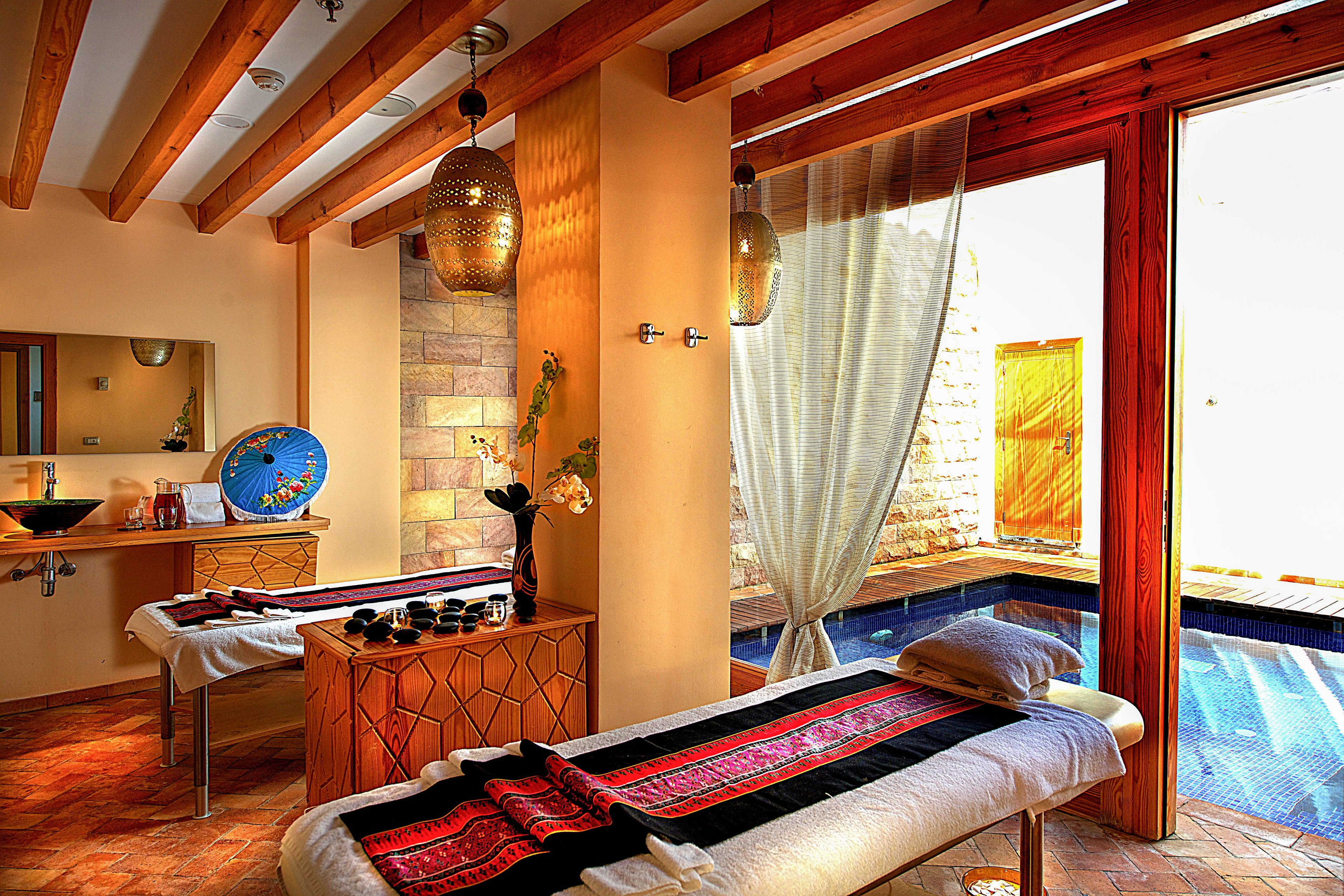 A serene spa treatment room features two massage tables adorned with patterned blankets and towels. The space is warmly lit with hanging lamps, wooden beams, and a view of a private pool through large glass doors. Decorative elements include a blue ceramic basin and a small arrangement of spa oils, creating a relaxing and inviting atmosphere. The earthy color palette and natural light enhance the tranquil mood.