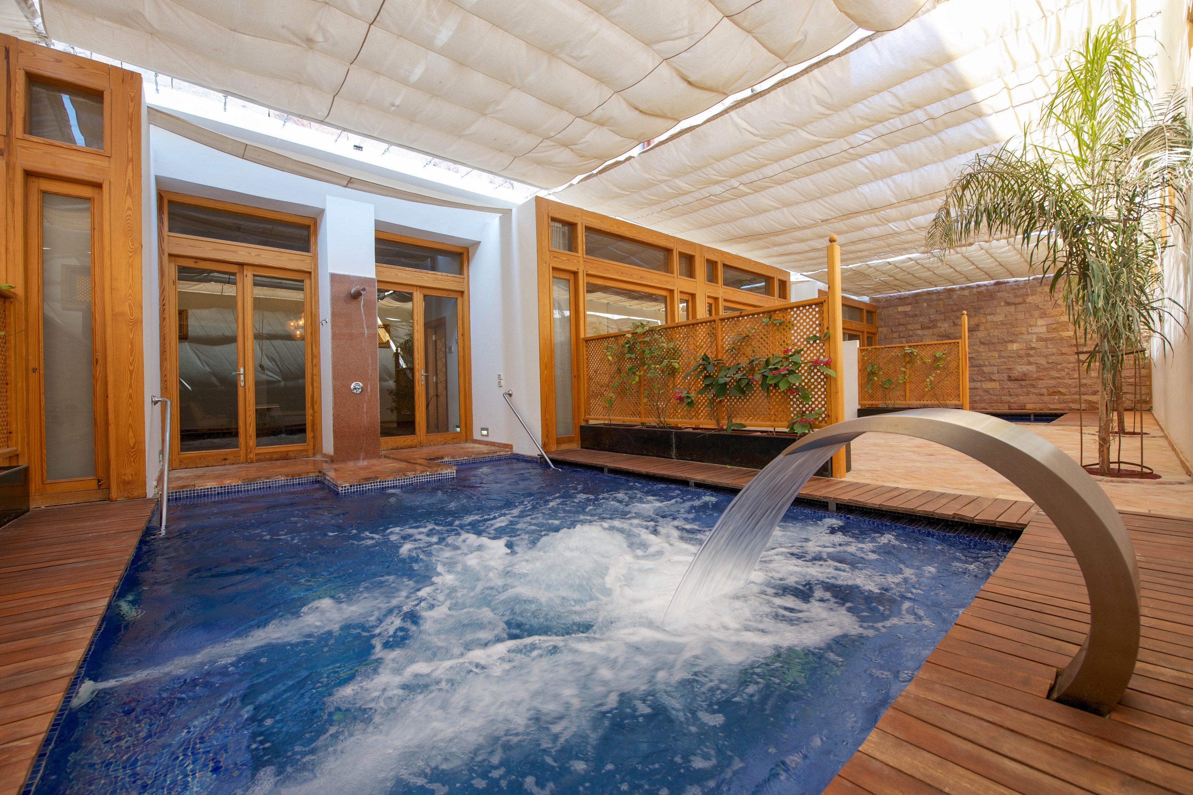 A contemporary indoor spa pool features bubbling water and a sleek metallic fountain. The setting includes a wooden deck, large glass doors, and a partially covered ceiling, creating a relaxing and luxurious atmosphere. Natural light filters through the translucent roof, and potted plants add a touch of greenery to the tranquil space.
