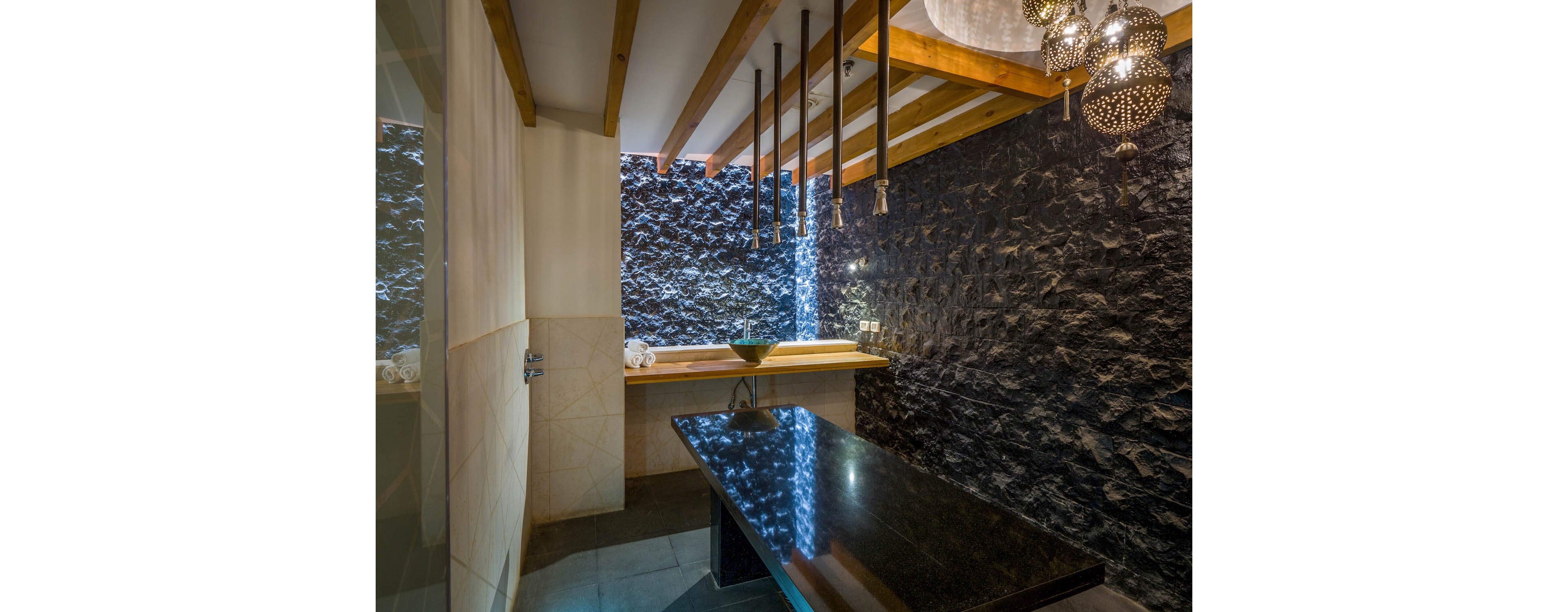A contemporary spa treatment room features a sleek black stone accent wall and a polished black massage table. The space is illuminated by hanging pendant lights and natural wood ceiling beams, creating a tranquil and luxurious atmosphere. A minimalist sink area with a wooden countertop adds to the modern aesthetic. The room is designed for relaxation and wellness treatments, with a focus on clean lines and calming textures.