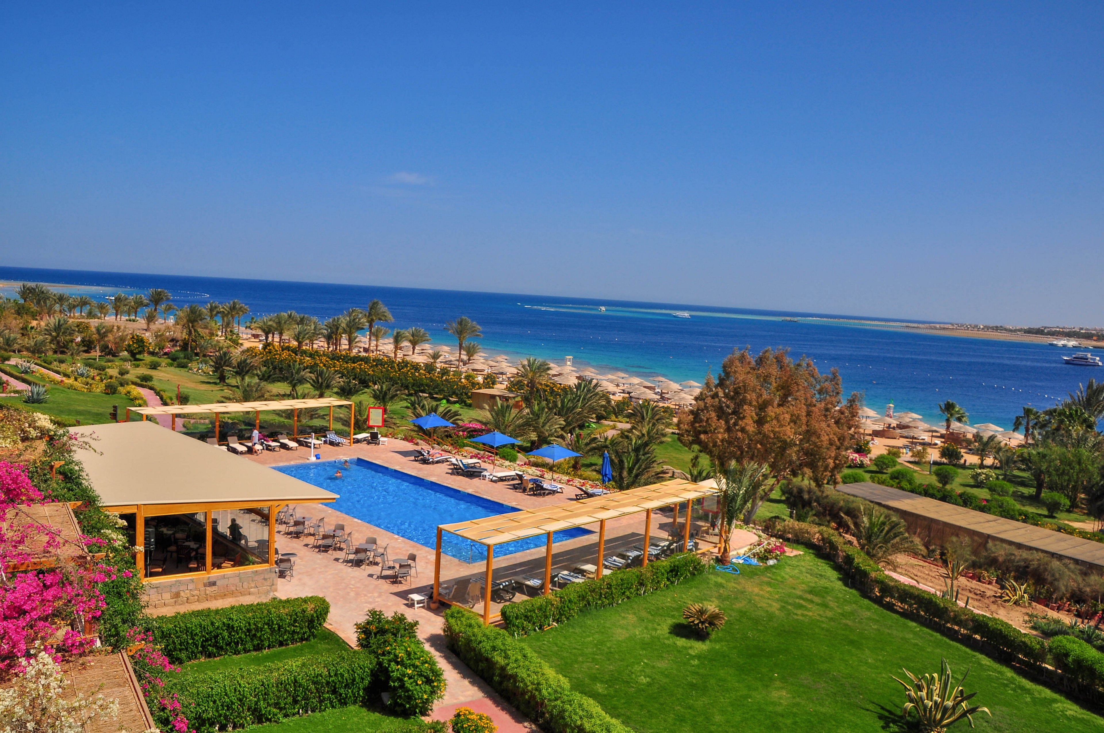 A vibrant resort features a large outdoor swimming pool surrounded by sun loungers and shaded pergolas. The setting includes lush green gardens and colorful flowers, with a sandy beach and clear blue sea in the background. The scene is bright and sunny, evoking a relaxing and inviting vacation atmosphere.