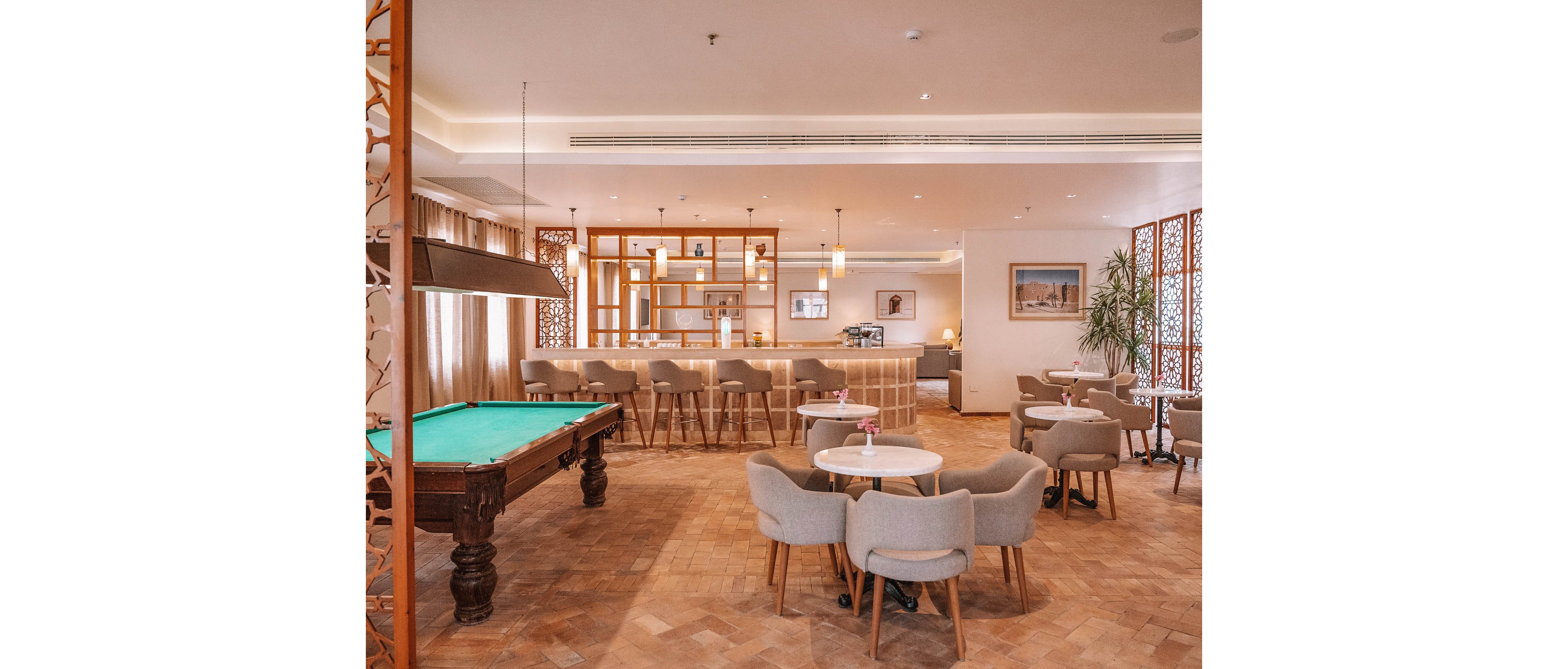 A contemporary lounge bar features a green pool table in the foreground and a stylish bar counter with high stools in the background. The setting is warm and inviting, with neutral-toned chairs and round tables arranged for social gatherings. Soft lighting, decorative wood accents, and framed artwork enhance the relaxed, upscale atmosphere. No visible text or numeric values are present in the image.
