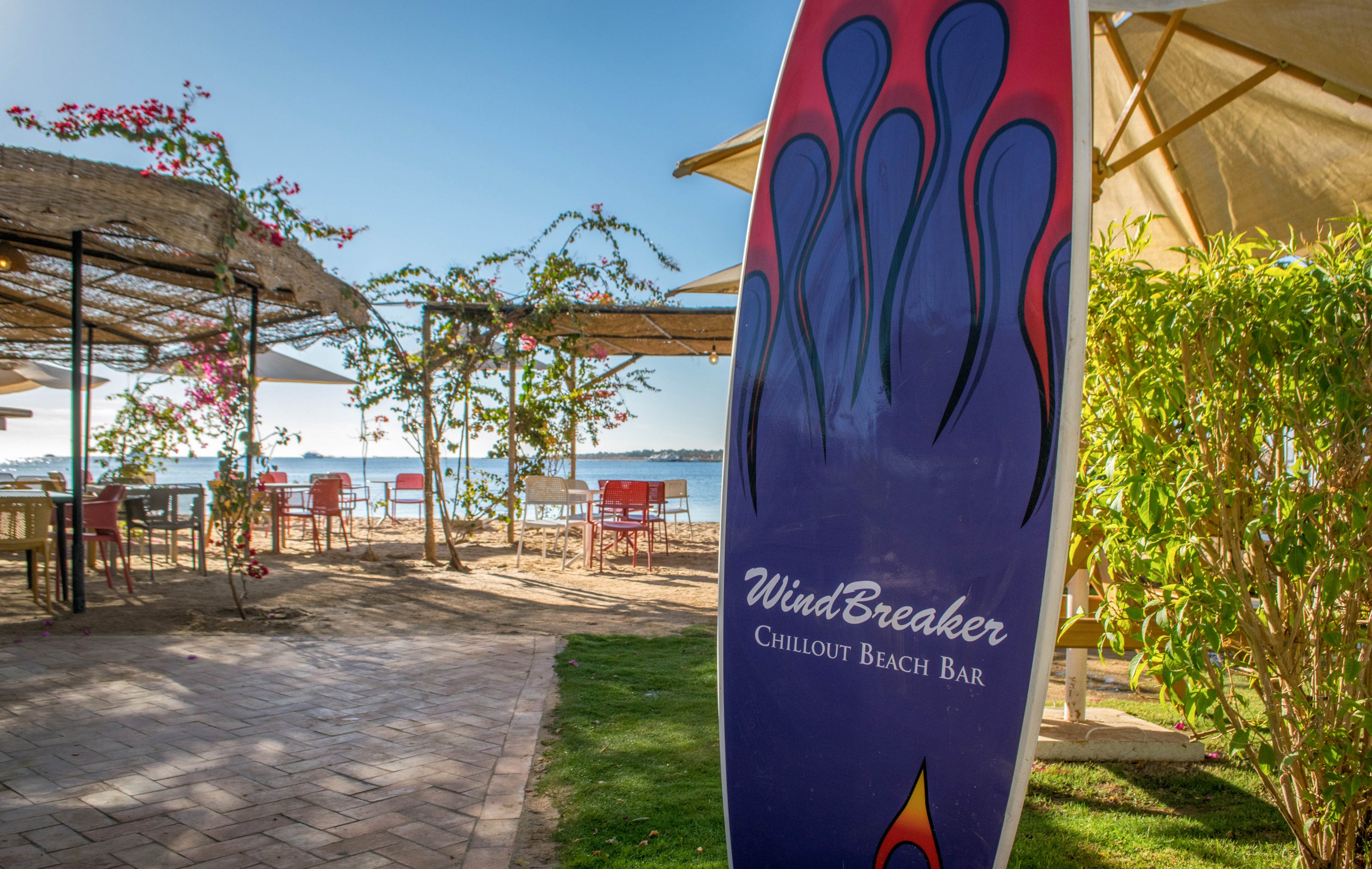 A colorful surfboard sign reading 'WindBreaker Chillout Beach Bar' stands prominently in the foreground of a sunny outdoor beach bar setting. The area features shaded seating, flowering vines, and a view of the sea in the background. The relaxed atmosphere is enhanced by natural light, greenery, and inviting patio furniture. The surfboard design adds a playful, coastal vibe to the scene.