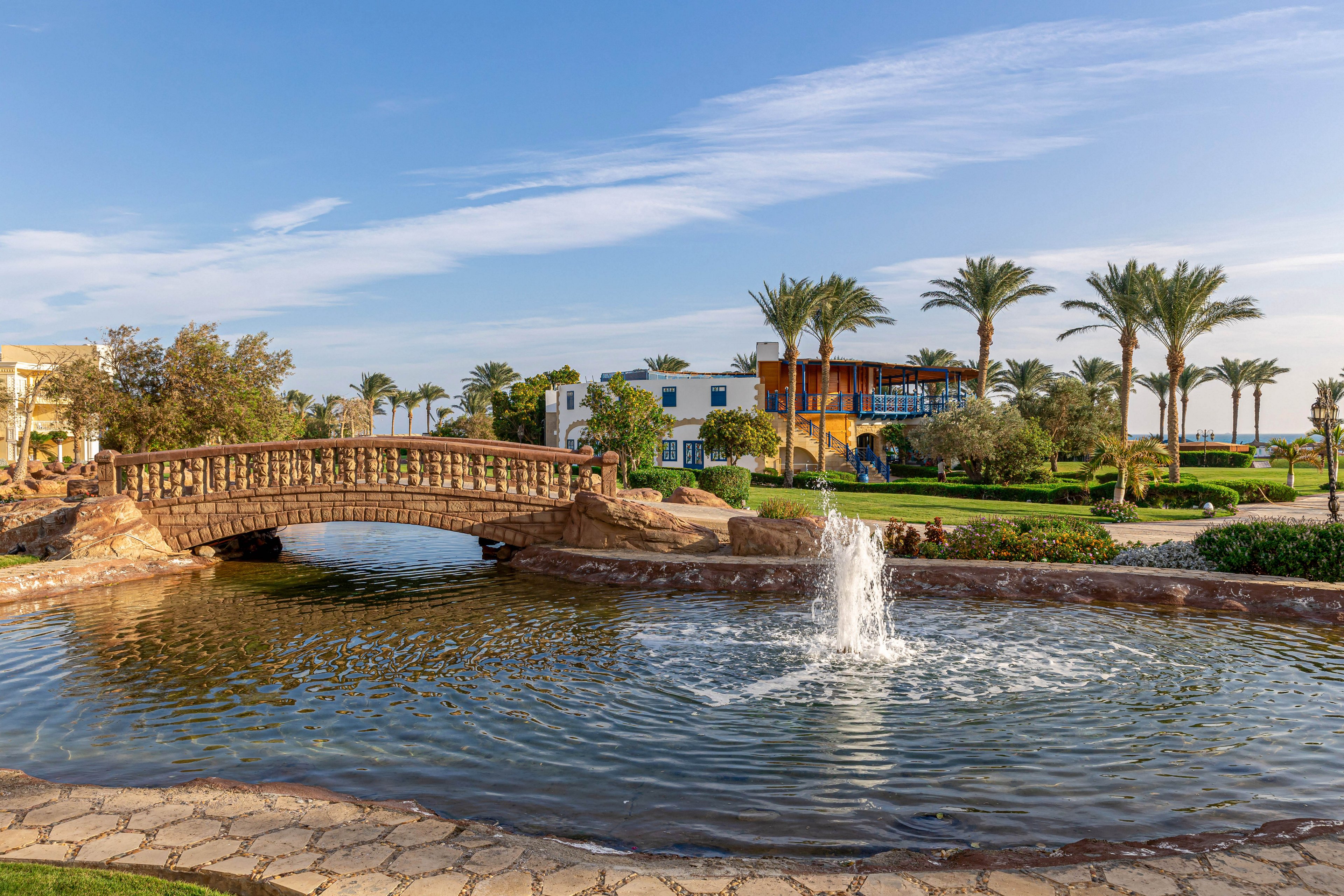 A picturesque stone bridge arches over a circular pond featuring a central water fountain. The scene is set in a landscaped resort garden with palm trees and manicured lawns, under a clear blue sky. Modern buildings are visible in the background, enhancing the tranquil atmosphere. The image conveys a peaceful, sunny mood with natural and architectural elements.