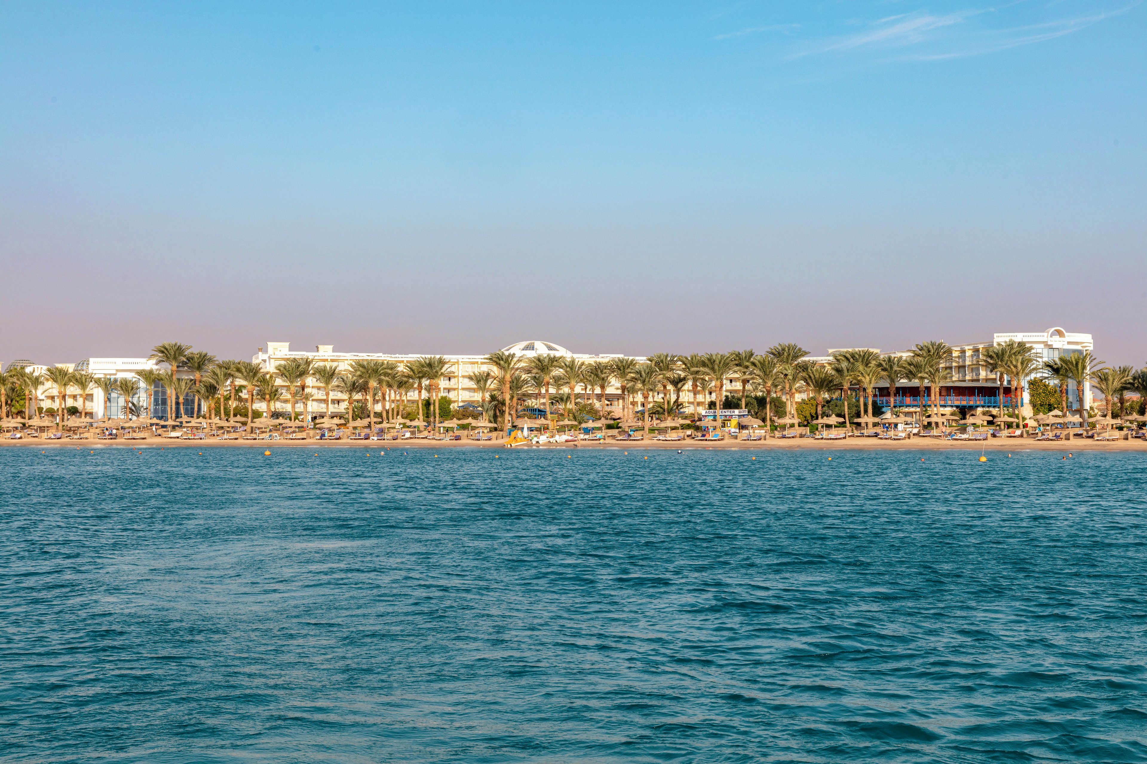 A large beachfront resort is visible in the distance, lined with rows of tall palm trees. The scene is set against a clear blue sky and calm sea, suggesting a tranquil and inviting atmosphere. The architecture is modern, with multiple buildings stretching along the shoreline. The mood is relaxed and summery, ideal for vacation or leisure activities. No visible text or numbers are present in the image.