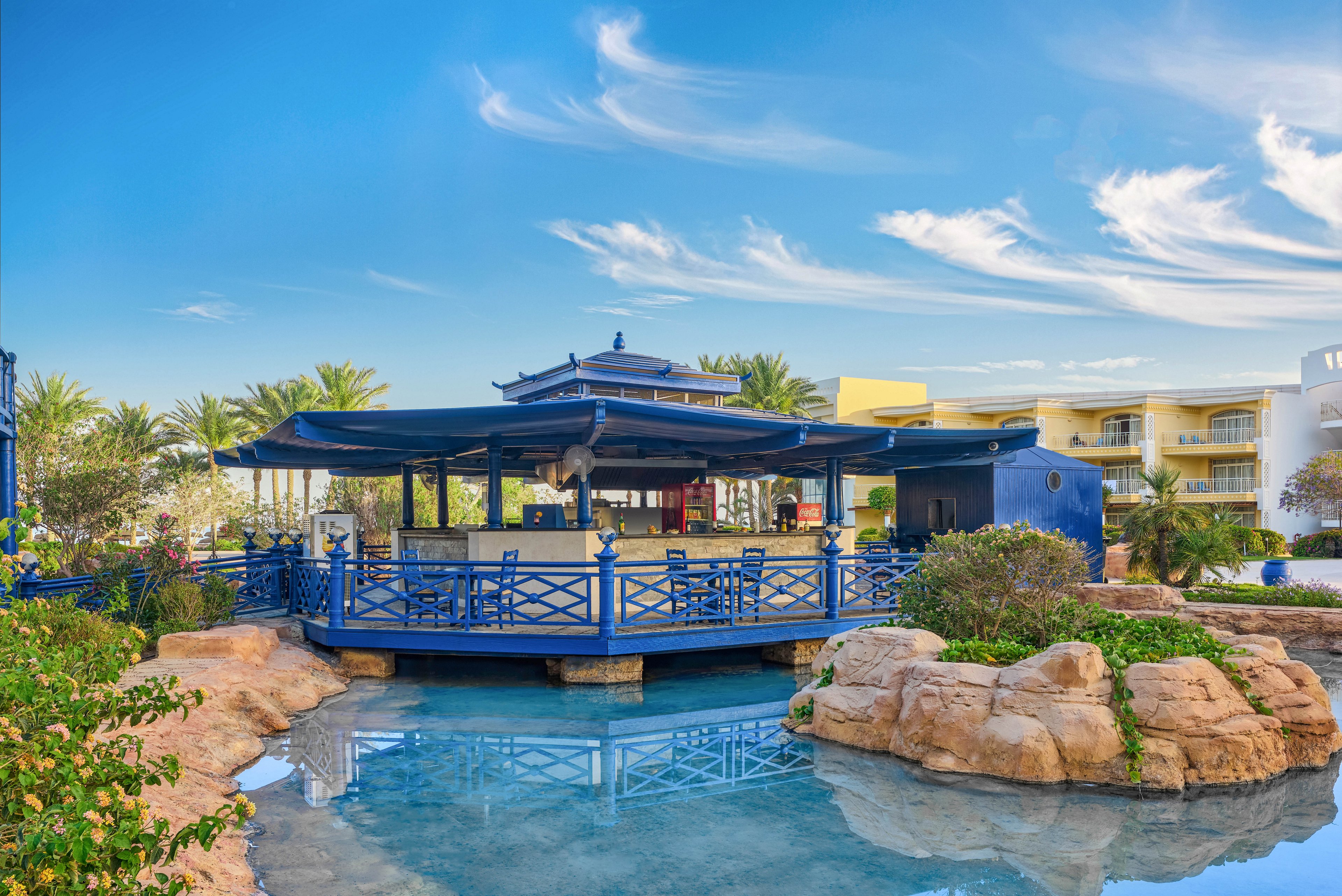 The main subject is a blue pavilion structure situated over a shallow water feature, surrounded by rocks and lush greenery. The setting appears to be a resort or hotel, with palm trees and modern buildings visible in the background. The scene is bright and tranquil, with clear skies and soft clouds enhancing the relaxed mood. No visible text or numeric values are present in the image.