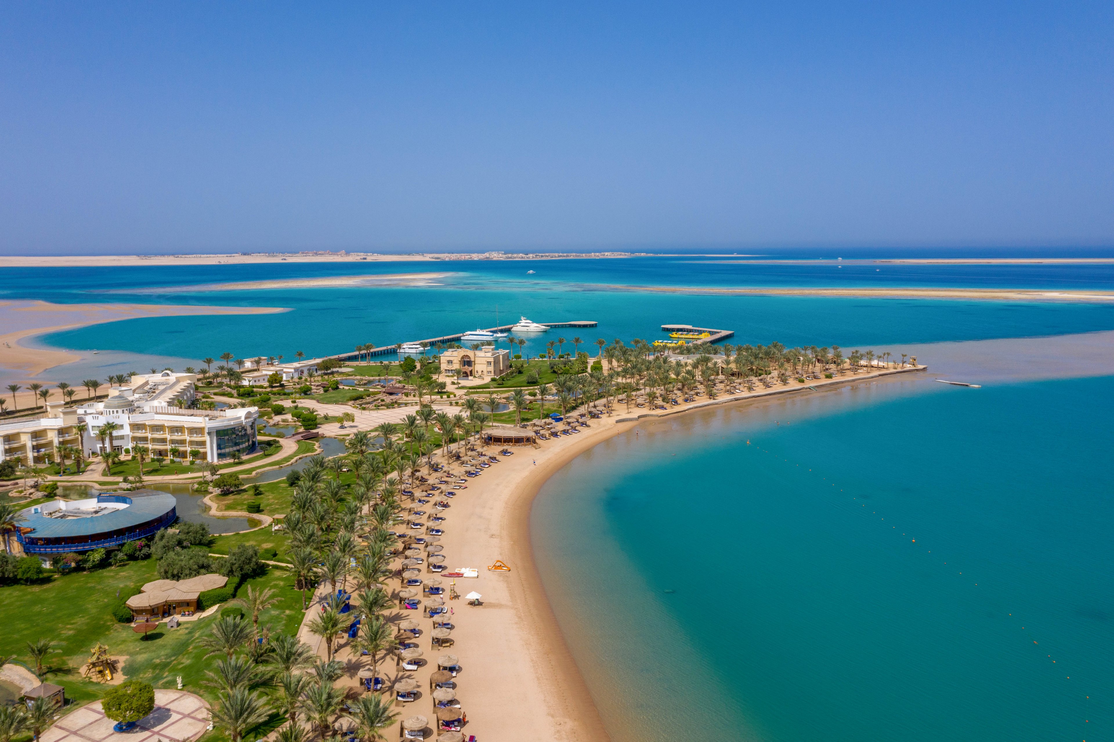The image shows a sweeping aerial perspective of a luxury beach resort with palm-lined shores and turquoise waters. Sun loungers and umbrellas are arranged along the sandy beach, while resort buildings and pools are visible in the background. The setting is tranquil, with clear blue skies and calm sea, evoking a relaxed vacation mood. No visible text or numeric values are present in the image.