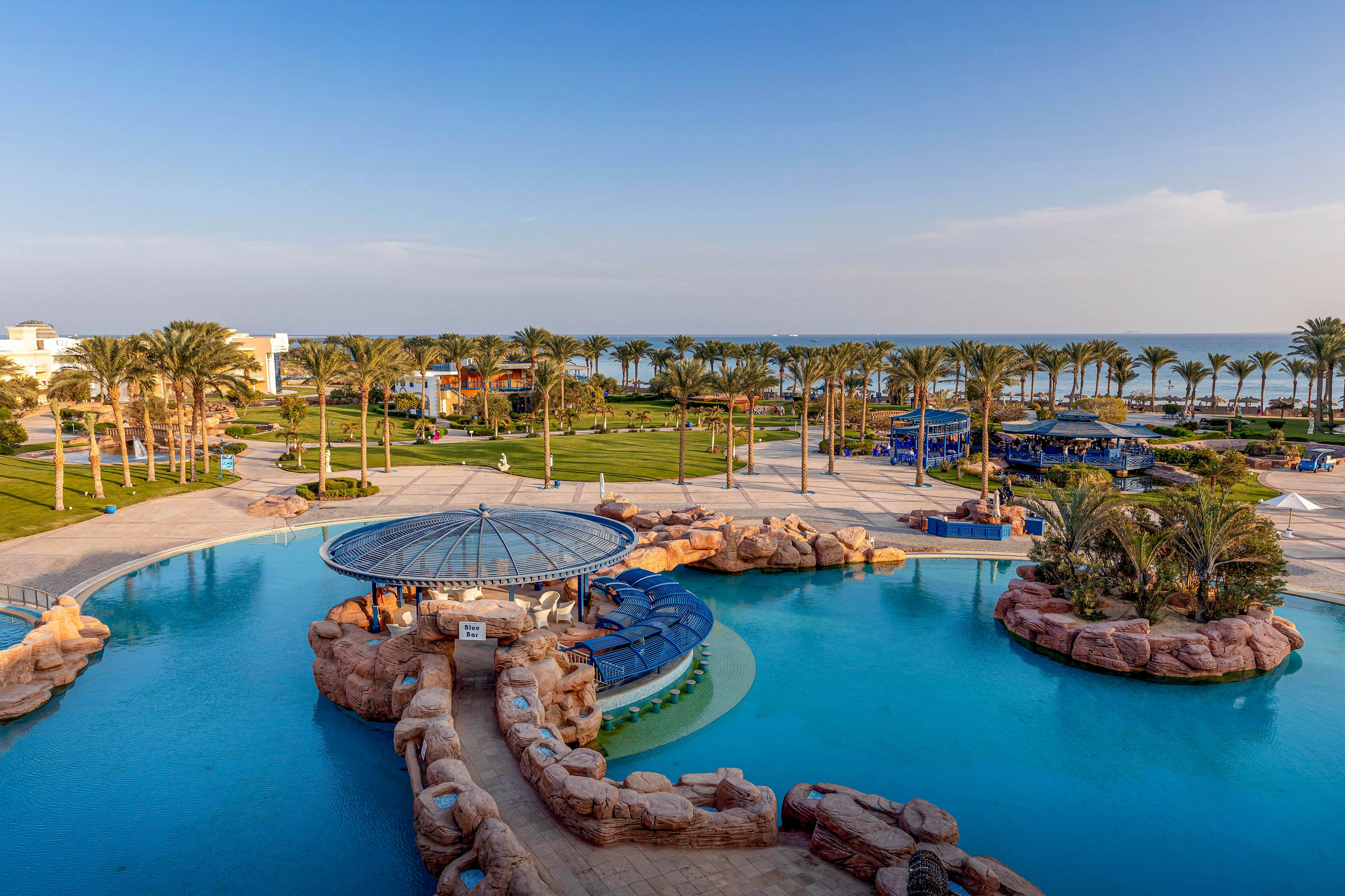 The image features a large, luxurious swimming pool with a circular swim-up bar surrounded by rock formations. Palm trees line the background, leading to a manicured lawn and the ocean beyond. The setting is tranquil and sunny, with clear blue skies and a relaxed vacation atmosphere. No visible text or numeric values are present in the image.