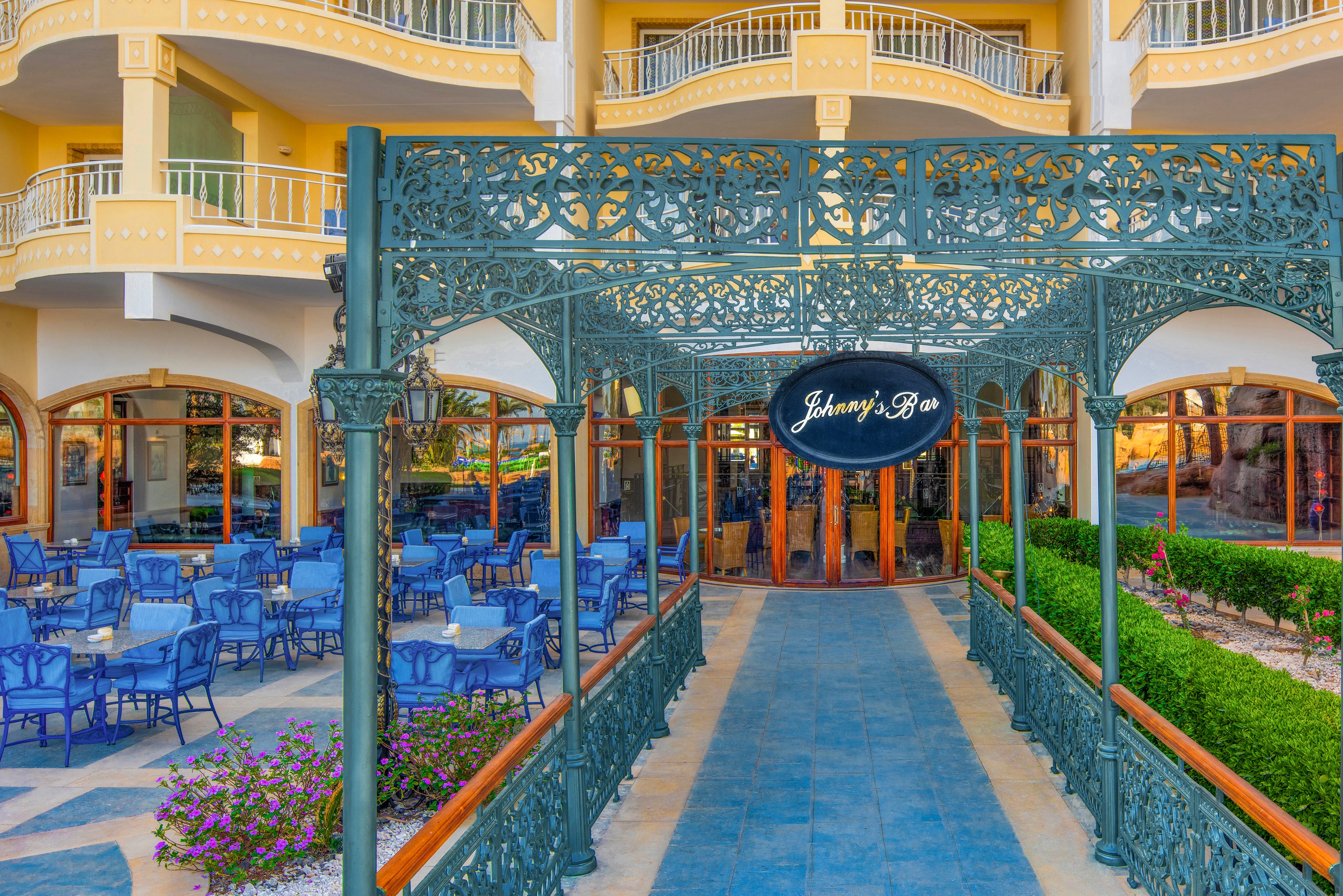 The image features the entrance to Johnny’s Bar, clearly marked by a black oval sign. The setting is an upscale hotel or resort with ornate ironwork and multiple balconies. Blue chairs and tables are arranged in the outdoor seating area, surrounded by manicured greenery and colorful flowers. The mood is vibrant and inviting, with warm yellow tones and a Mediterranean architectural style.
