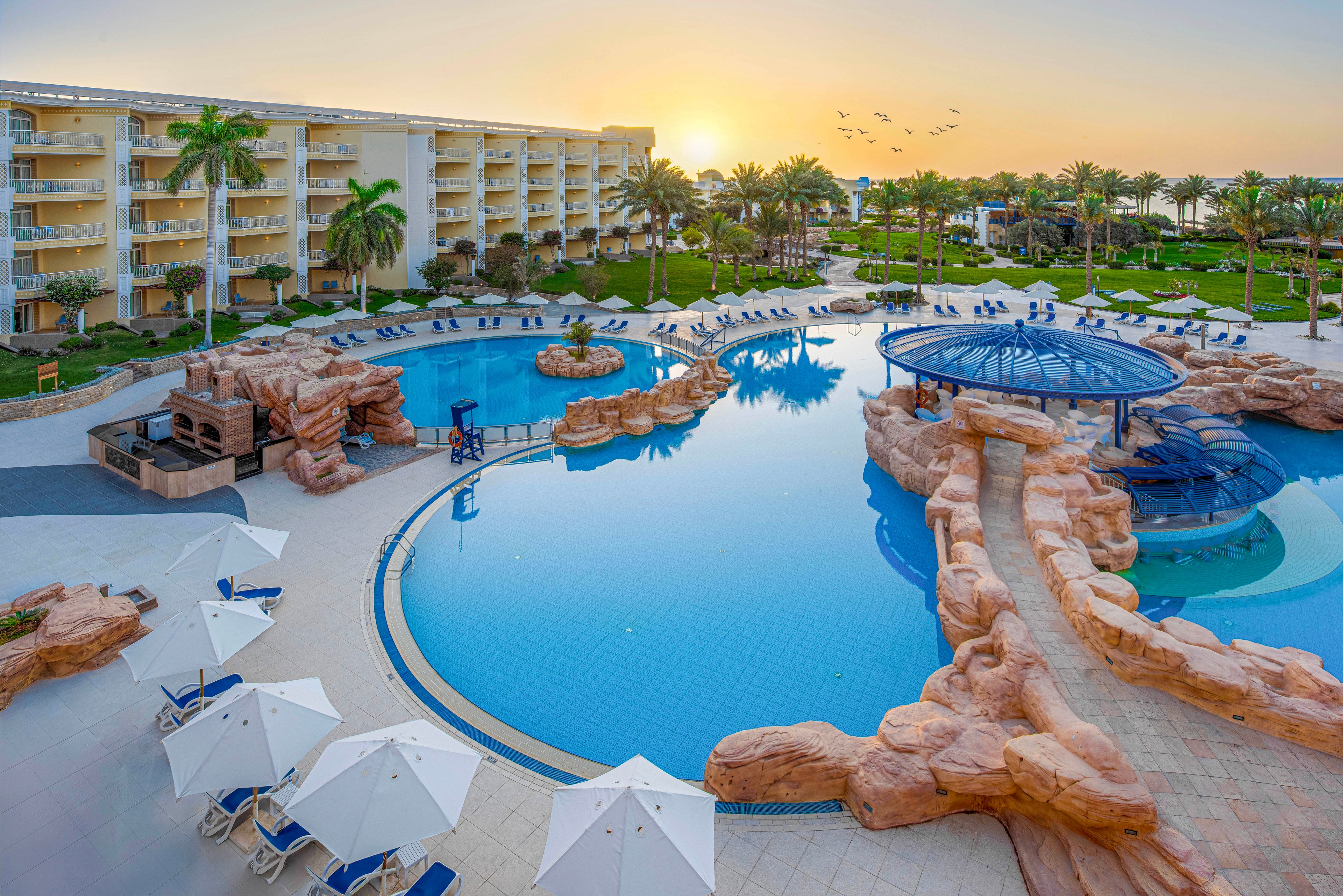 A large, lagoon-style swimming pool is the centerpiece of this luxury resort, surrounded by sun loungers and white umbrellas. The setting features lush palm trees, manicured lawns, and a multi-story hotel building in the background. The tranquil scene is captured at sunrise, with soft golden light and a flock of birds flying overhead. The pool includes rock formations and a shaded circular seating area. No visible text or numbers are present in the image.