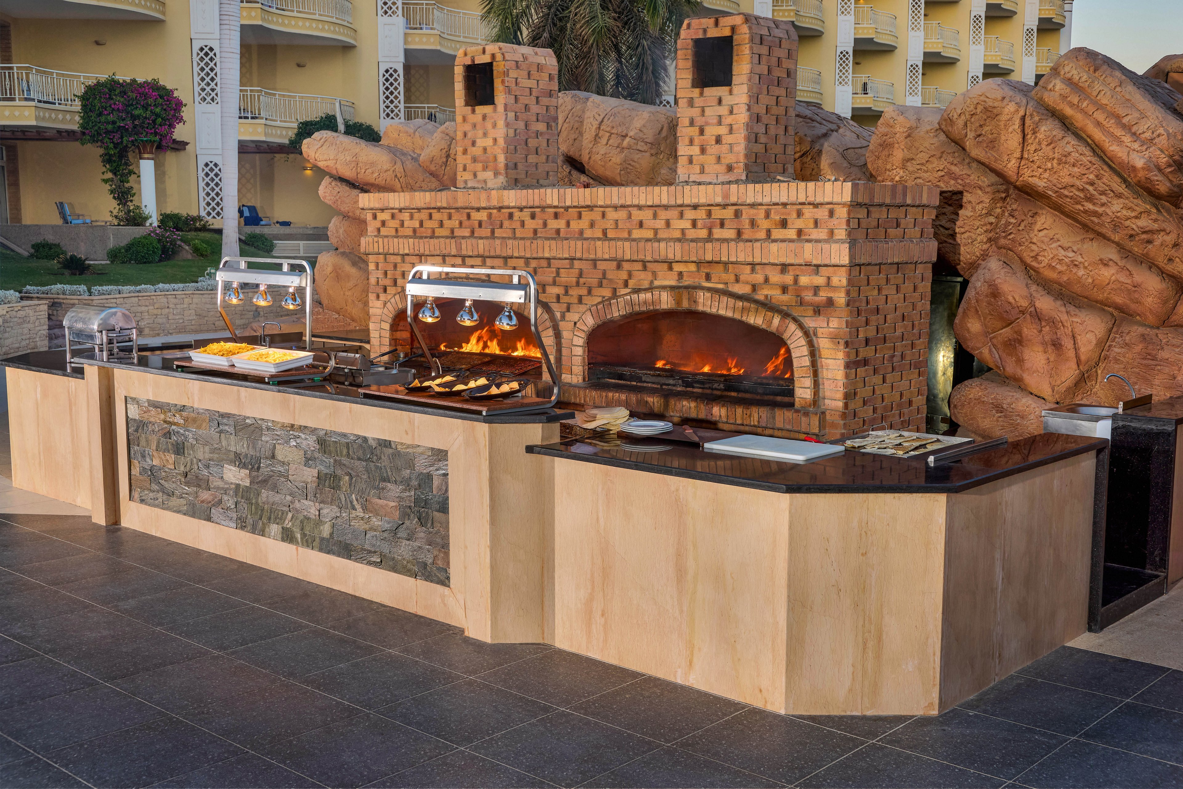 Two chefs work at an outdoor kitchen featuring a large brick oven and modern serving stations. The setting appears to be a resort or hotel terrace, with a backdrop of balconies and lush landscaping. The kitchen counter is made of stone and wood, and the oven is actively in use with visible flames. Food is displayed under heat lamps, and the area is clean and organized. No visible text or numbers are present in the image.