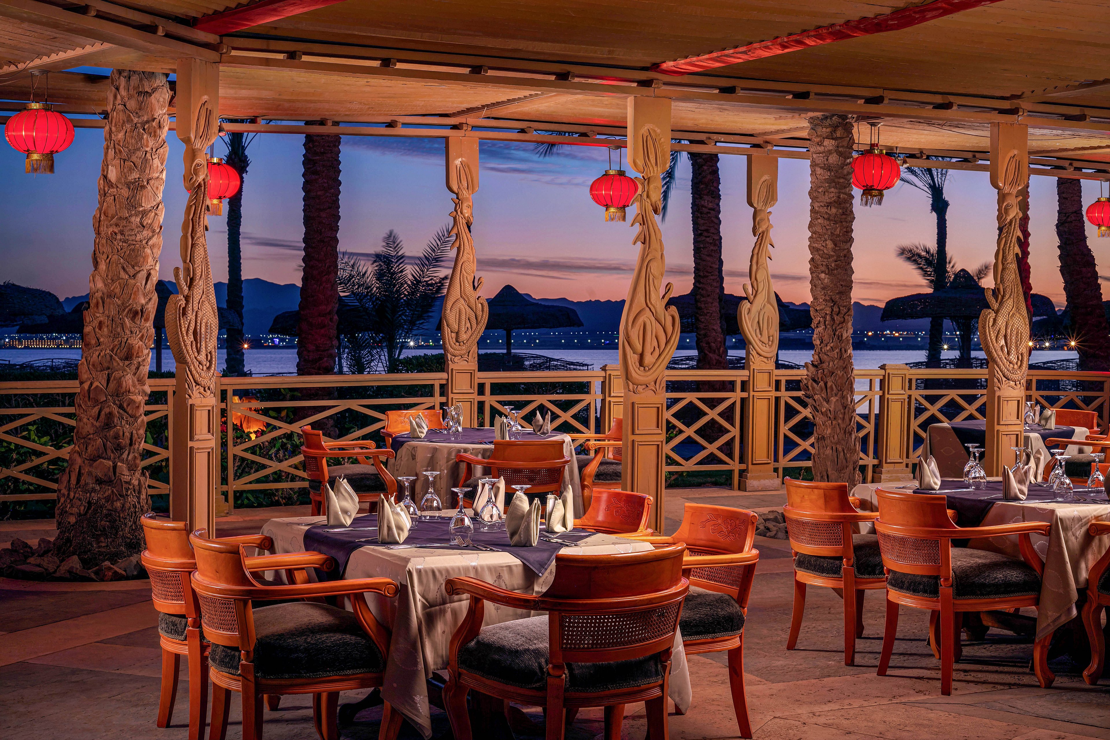 The image shows an elegant outdoor restaurant setup with wooden chairs and tables, each arranged with folded napkins and glassware. Red lanterns hang from the ceiling, casting a warm glow over the scene. The setting overlooks a scenic waterfront at dusk, with palm trees and distant mountains visible. The mood is tranquil and inviting, ideal for evening dining.