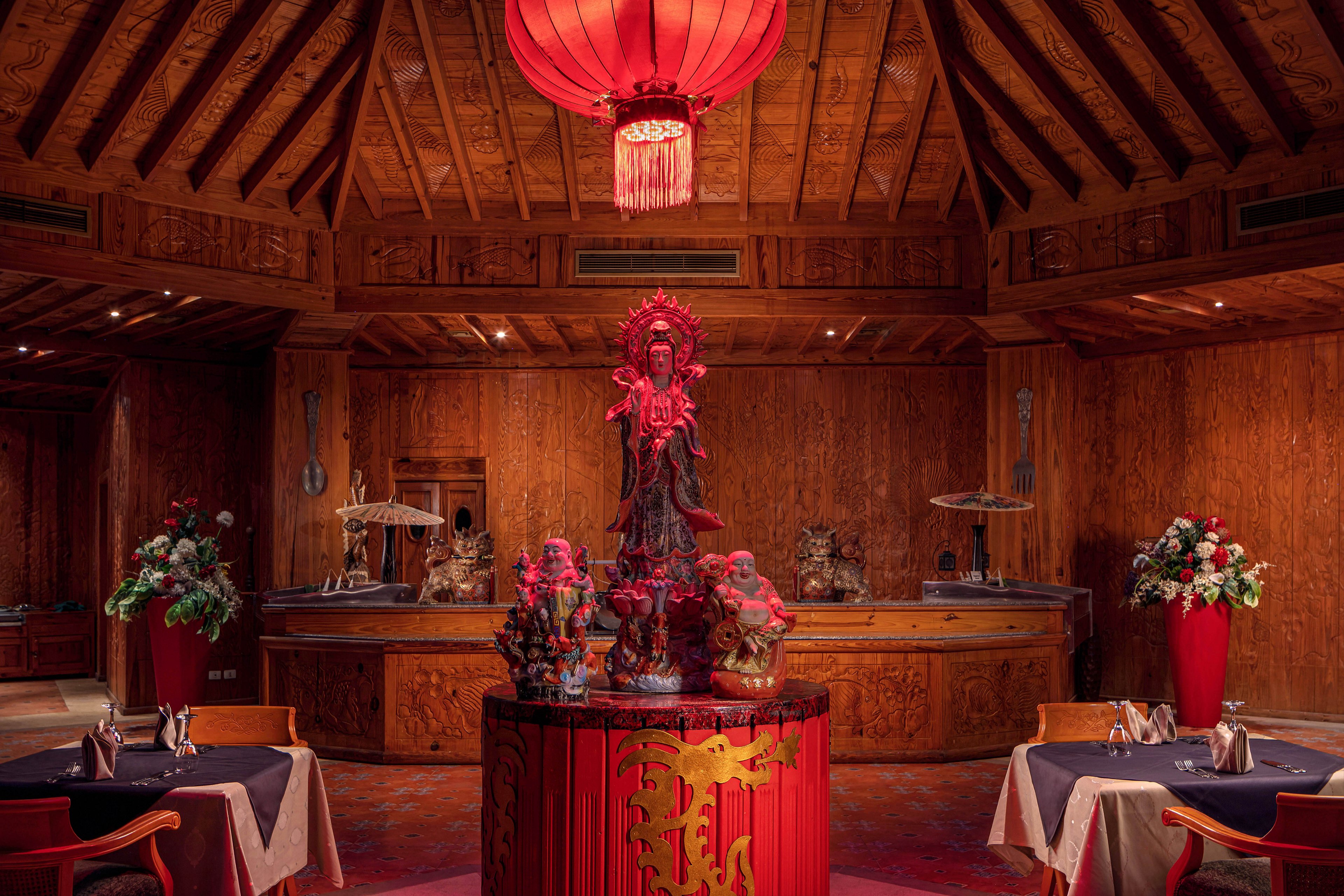 The main subject is a striking statue centerpiece surrounded by ornate decorations in a wood-paneled dining room. The setting features round tables with dark tablecloths, red chairs, and floral arrangements in tall red vases. A large red lantern hangs overhead, casting a warm glow and enhancing the rich, luxurious ambiance. The visuals highlight Asian motifs and intricate details throughout the space.