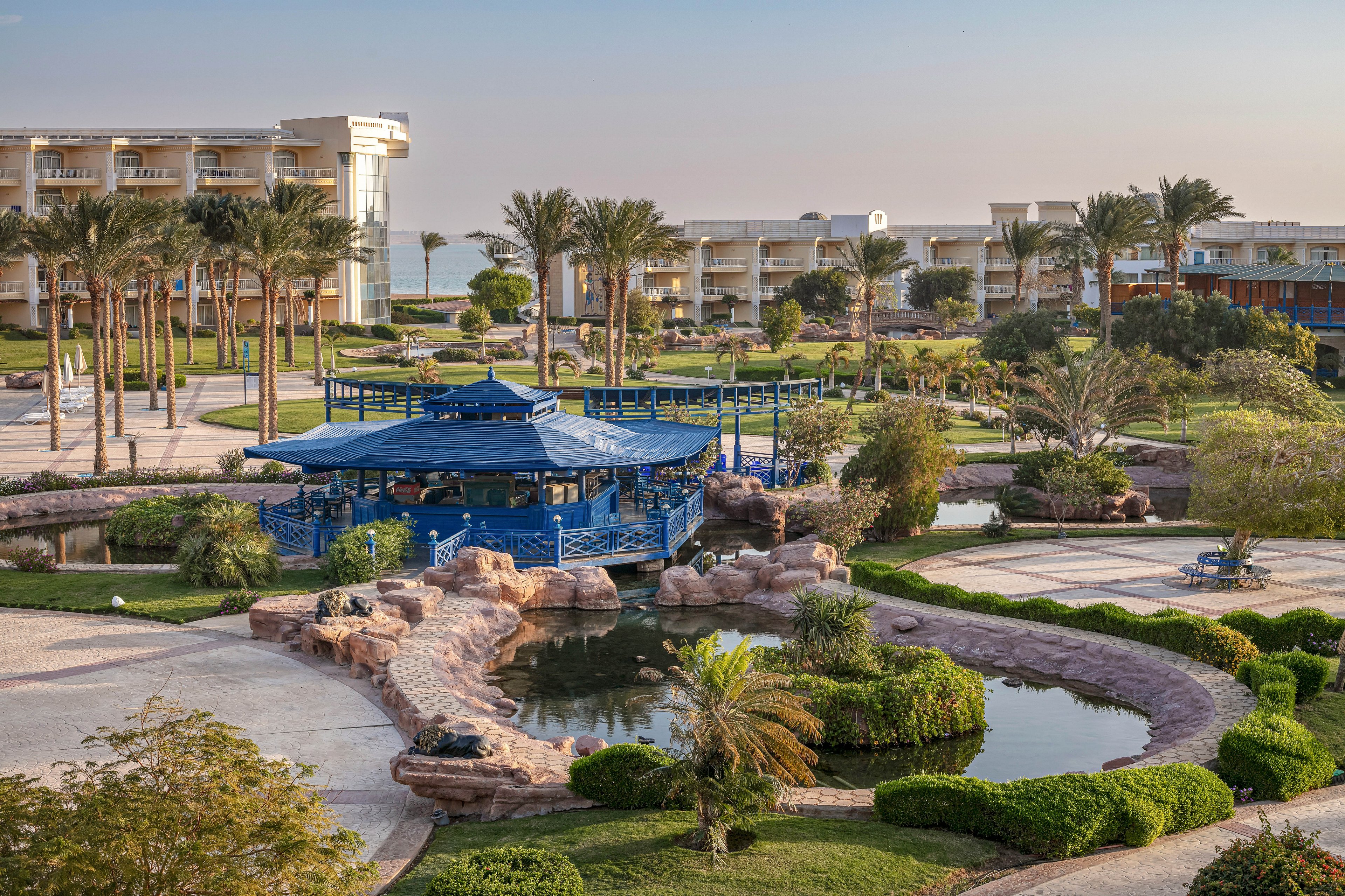 The image features a landscaped resort garden with a prominent blue pavilion surrounded by lush greenery and palm trees. Pathways wind through the area, connecting water features and manicured lawns. Modern hotel buildings are visible in the background, suggesting a tranquil vacation setting. The overall mood is serene and inviting, with soft natural lighting and a warm color palette.