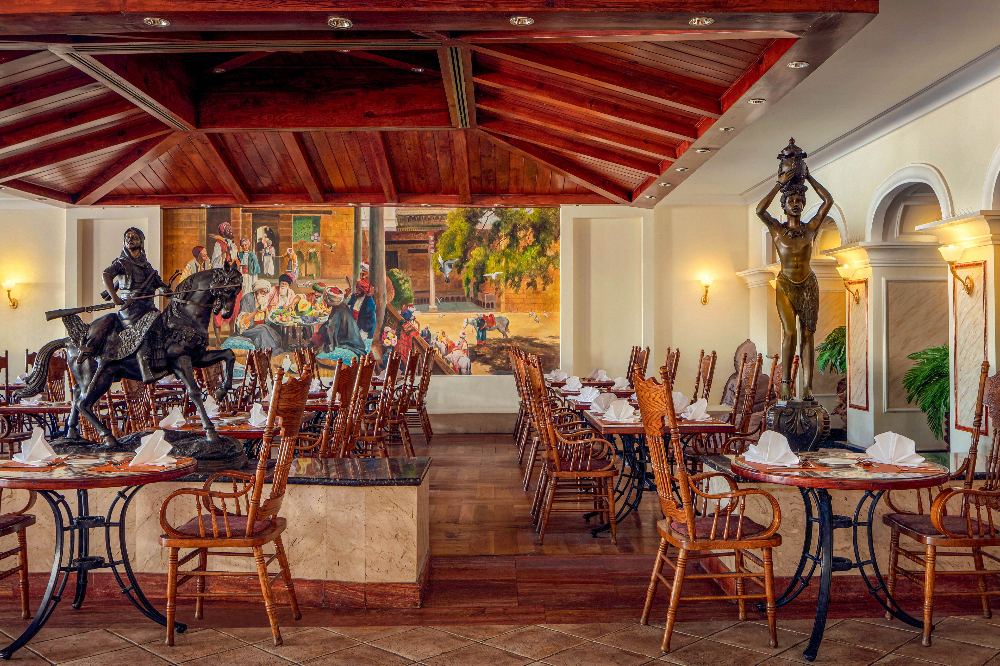 The image features a sophisticated restaurant dining area with wooden chairs and tables, each set with white napkins. Two prominent statues, one of a knight on horseback and another of a standing figure, are visible near the entrance. A vibrant mural depicting a lively historical scene adorns the back wall, adding color and visual interest. Warm lighting and rich wood tones create a welcoming and upscale atmosphere.