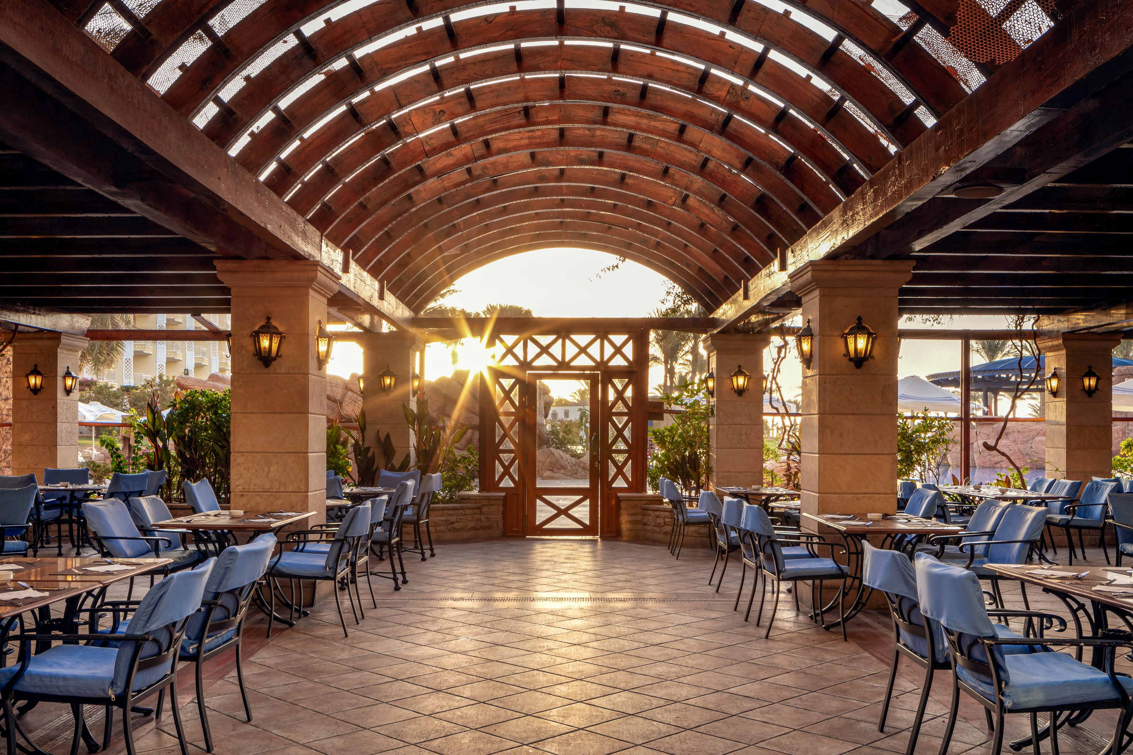The image shows an open-air restaurant patio featuring a striking arched wooden roof structure. Rows of empty tables and blue chairs are neatly arranged, creating a welcoming dining atmosphere. Warm sunlight streams through the latticework, casting soft shadows and highlighting the tiled floor. Lantern-style wall lights add a cozy touch, while lush greenery surrounds the space. No visible text or numbers are present in the scene.
