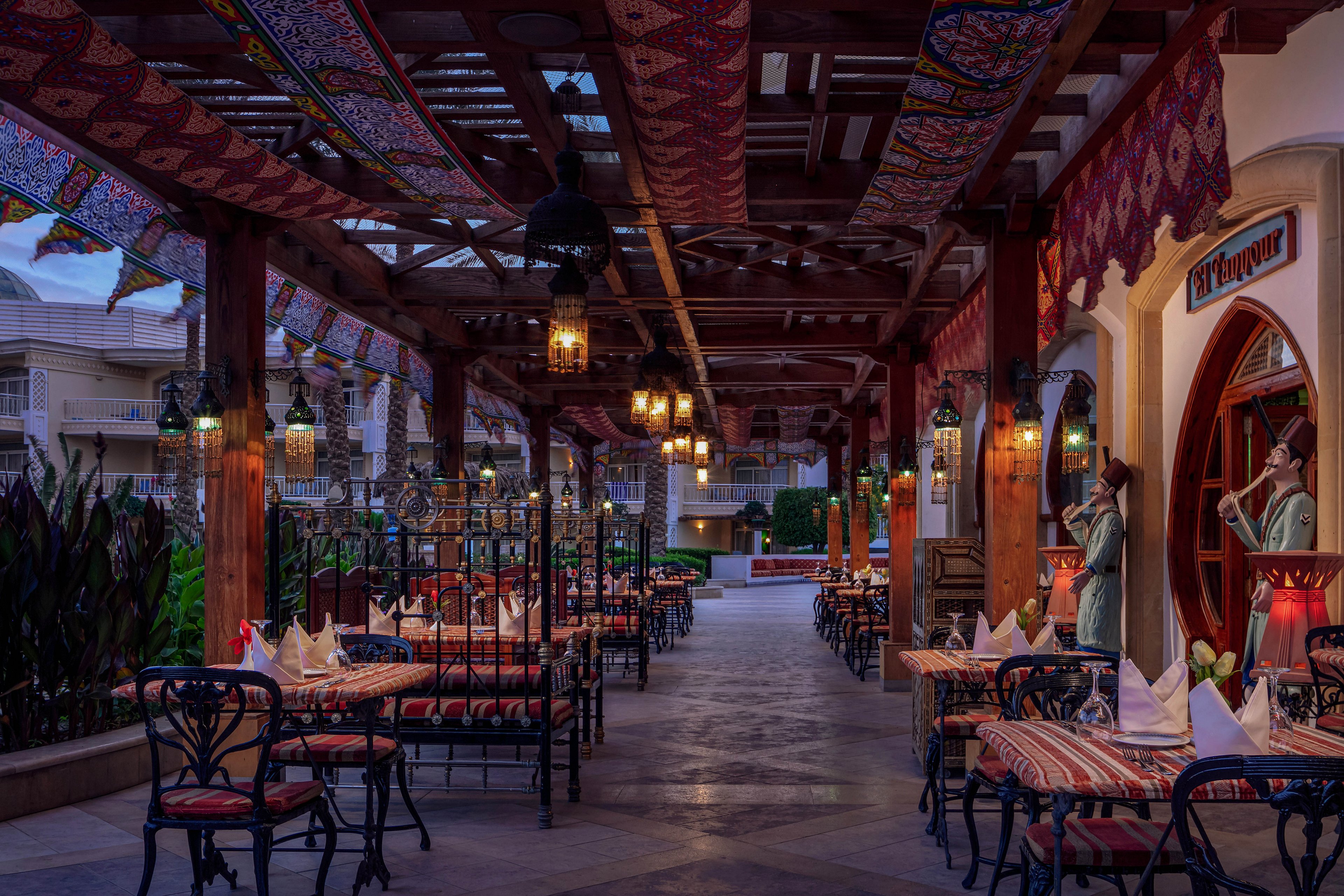 The image shows an inviting outdoor restaurant terrace, featuring tables set with folded napkins and decorative tablecloths. The setting is enhanced by hanging lanterns and colorful patterned fabrics draped overhead, creating a warm and cozy ambiance. Two statues of musicians are visible near the entrance, adding a cultural touch. The lighting and evening atmosphere contribute to a relaxed, elegant mood.