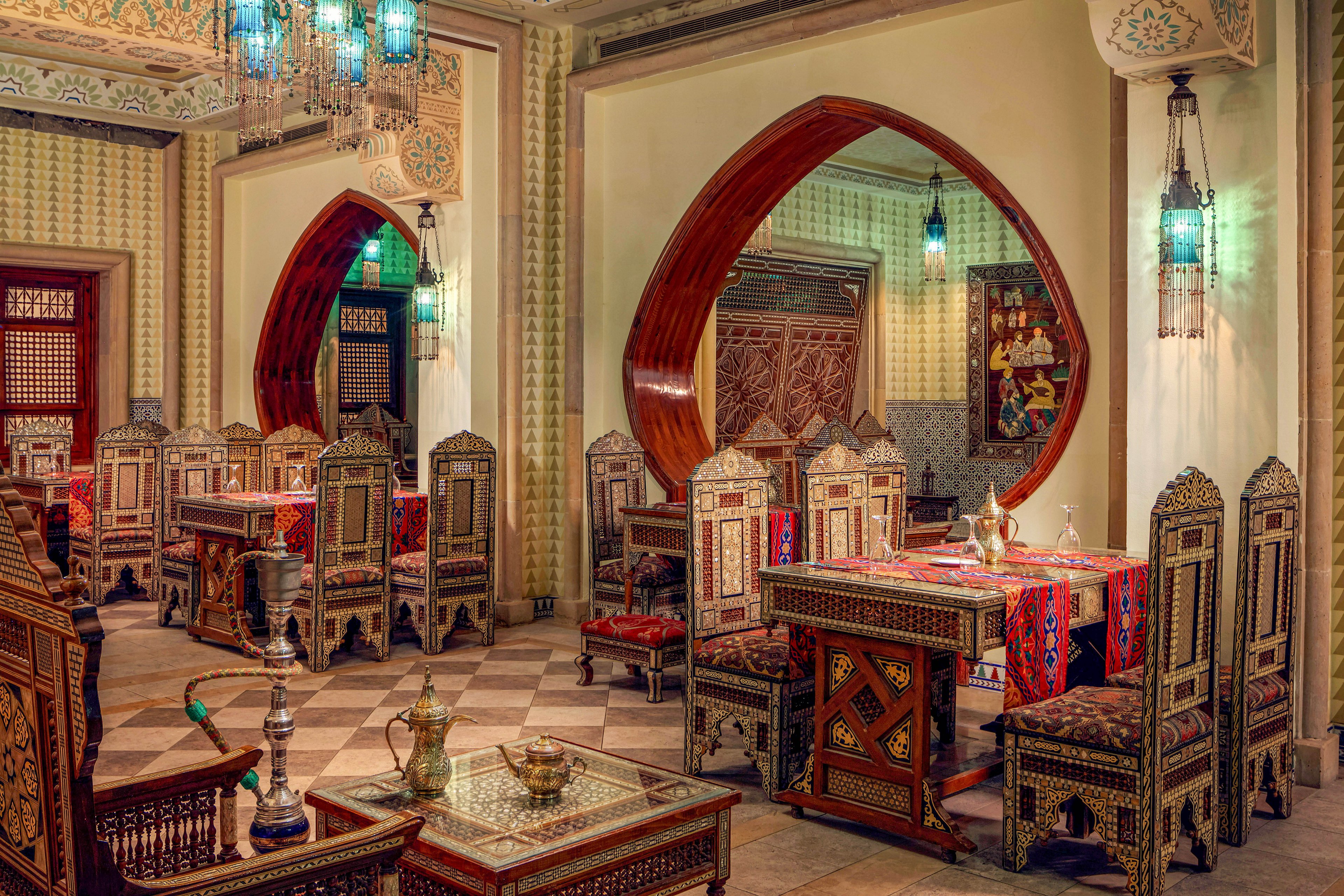 This image features a richly decorated Middle Eastern restaurant interior with intricately carved wooden chairs and tables. The setting includes patterned textiles, hanging lanterns with blue-green light, and a hookah placed on a side table. The arched wooden frames and mosaic tilework enhance the authentic ambiance, while the absence of people highlights the detailed craftsmanship and inviting atmosphere.