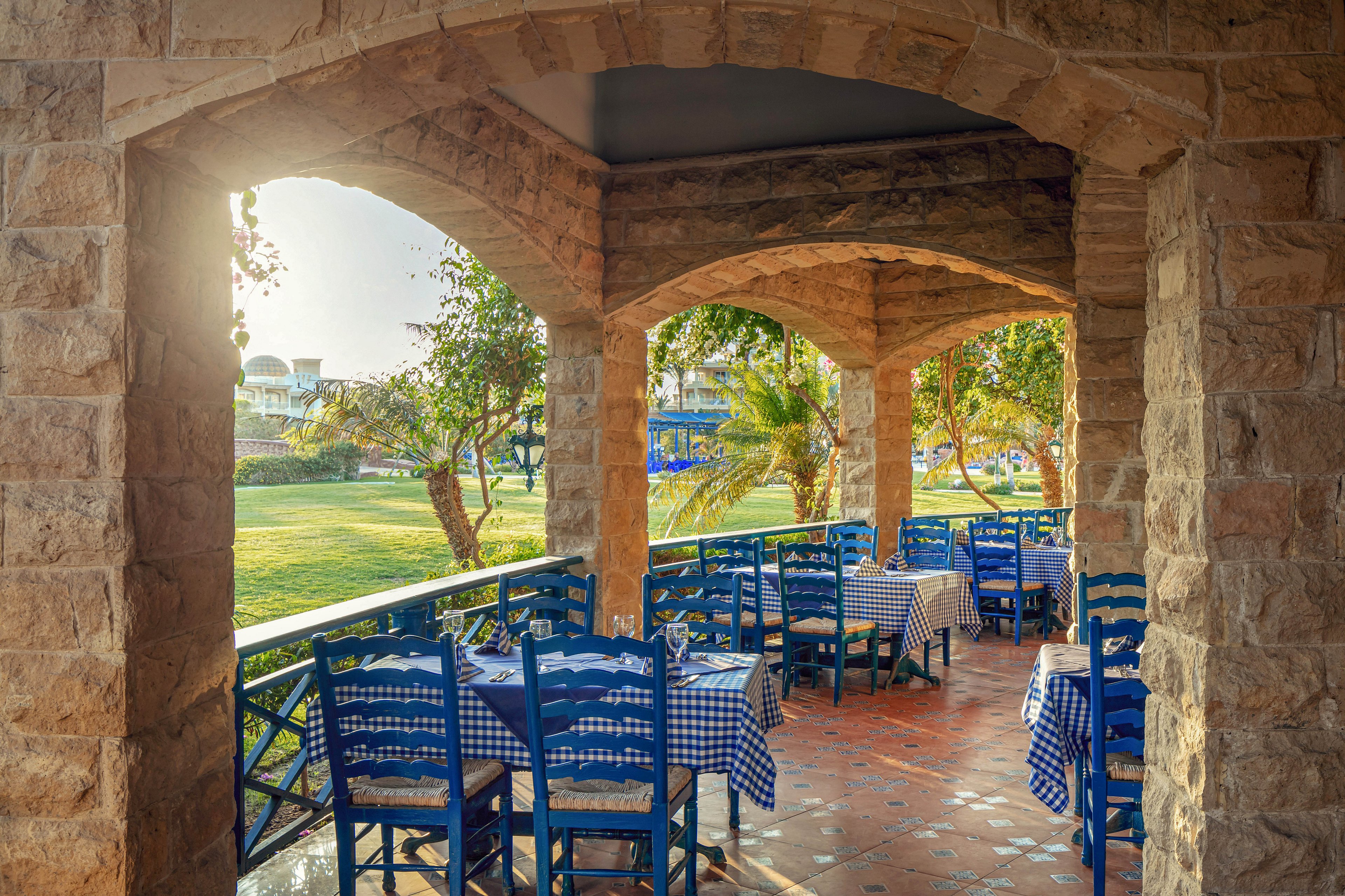 The image shows a charming outdoor dining area with blue wooden chairs and tables covered in blue and white checkered cloths. The terrace is framed by stone arches, overlooking a lush green garden with palm trees. Sunlight streams through the arches, creating a warm and inviting atmosphere. No visible text or numeric values are present in the scene.