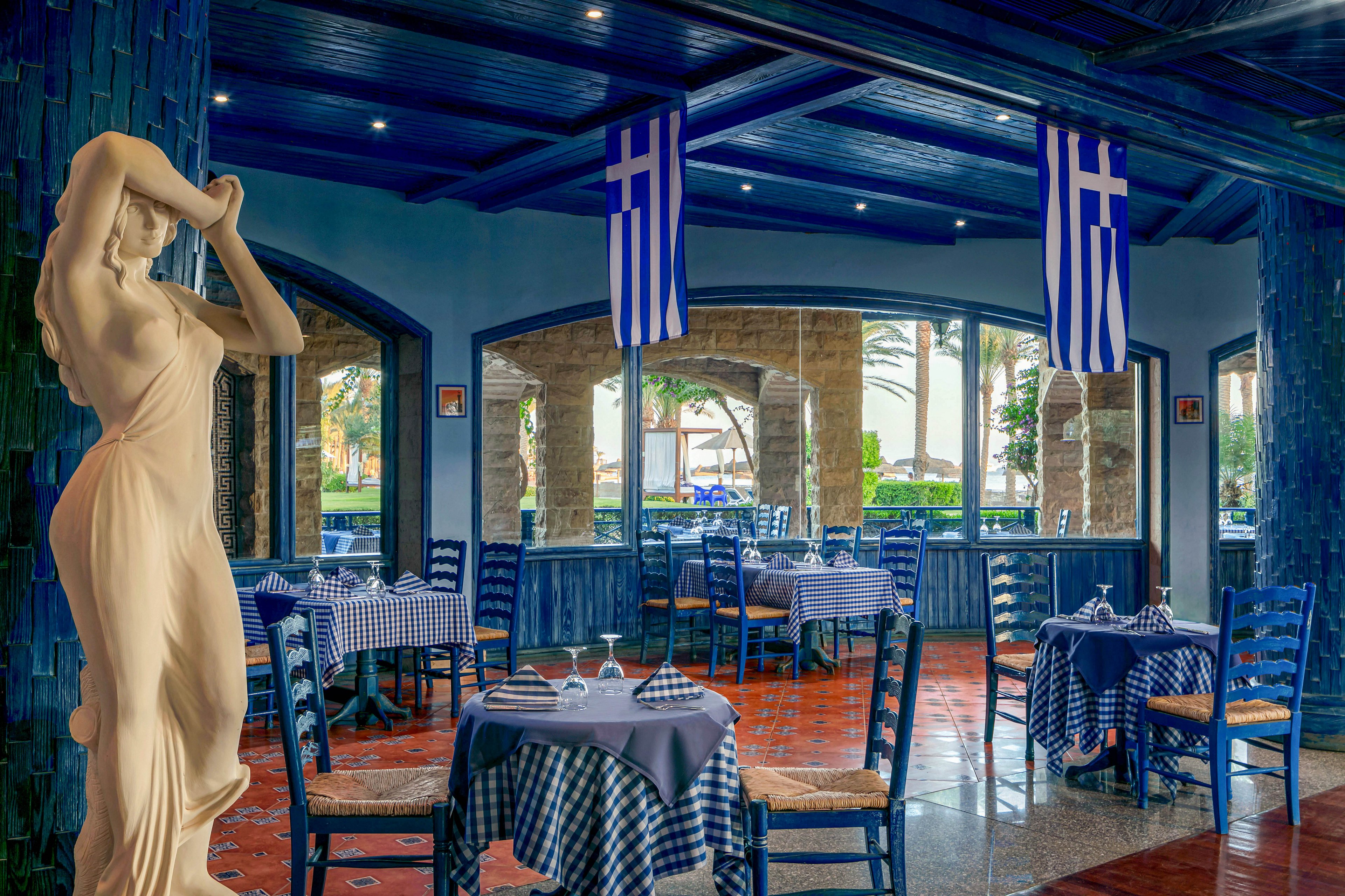 The image shows a Greek-style restaurant interior featuring blue decor, checkered tablecloths, and a classical statue. Greek flags hang from the ceiling, reinforcing the cultural theme. Large windows provide natural light and a view of palm trees outside. The setting is calm and inviting, with neatly arranged tables and chairs.