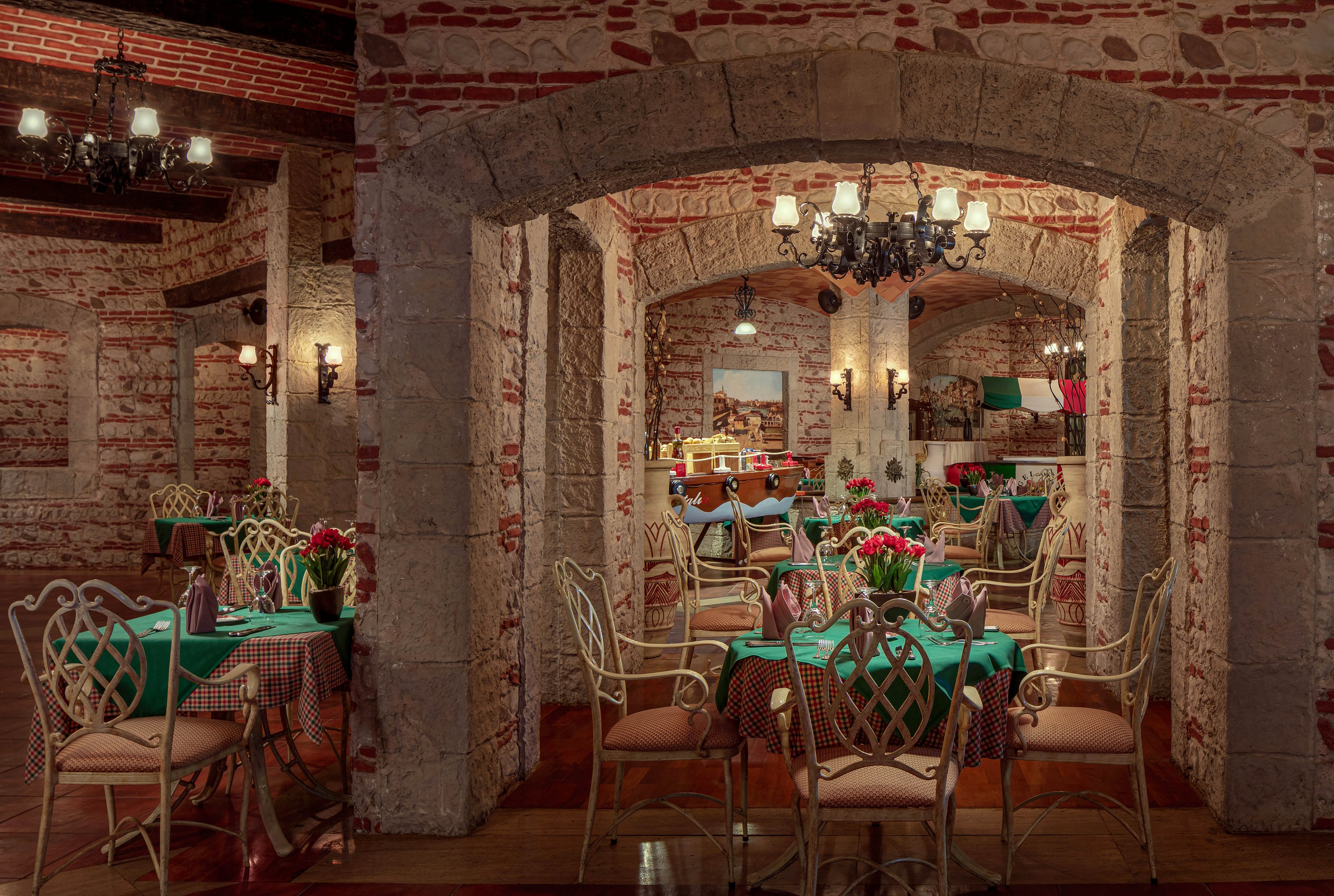 The image shows a warmly lit restaurant interior featuring exposed brick arches and elegant wrought iron chairs. Tables are set with green tablecloths and red floral centerpieces, creating a welcoming atmosphere. Chandeliers and wall sconces provide soft lighting, while the overall palette includes earthy tones and pops of red and green. The setting appears to be a traditional dining space, possibly Italian-themed, with a mural and flag visible in the background.