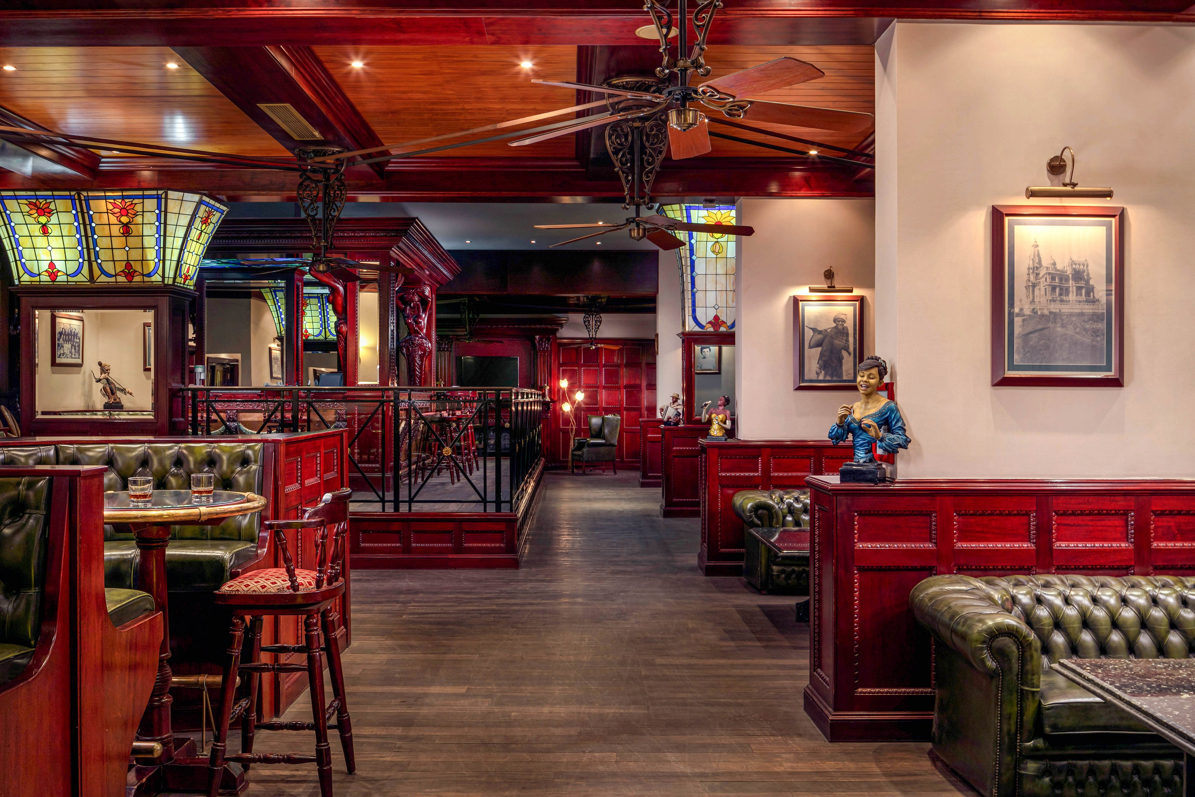 The image shows a traditional pub setting featuring dark wood paneling, green leather seating, and stained glass light fixtures. The space is empty, highlighting the cozy booths and ornate decor, including framed artwork and a decorative statue. Warm lighting and rich colors create an inviting, vintage atmosphere. No visible text or numeric values are present in the scene.