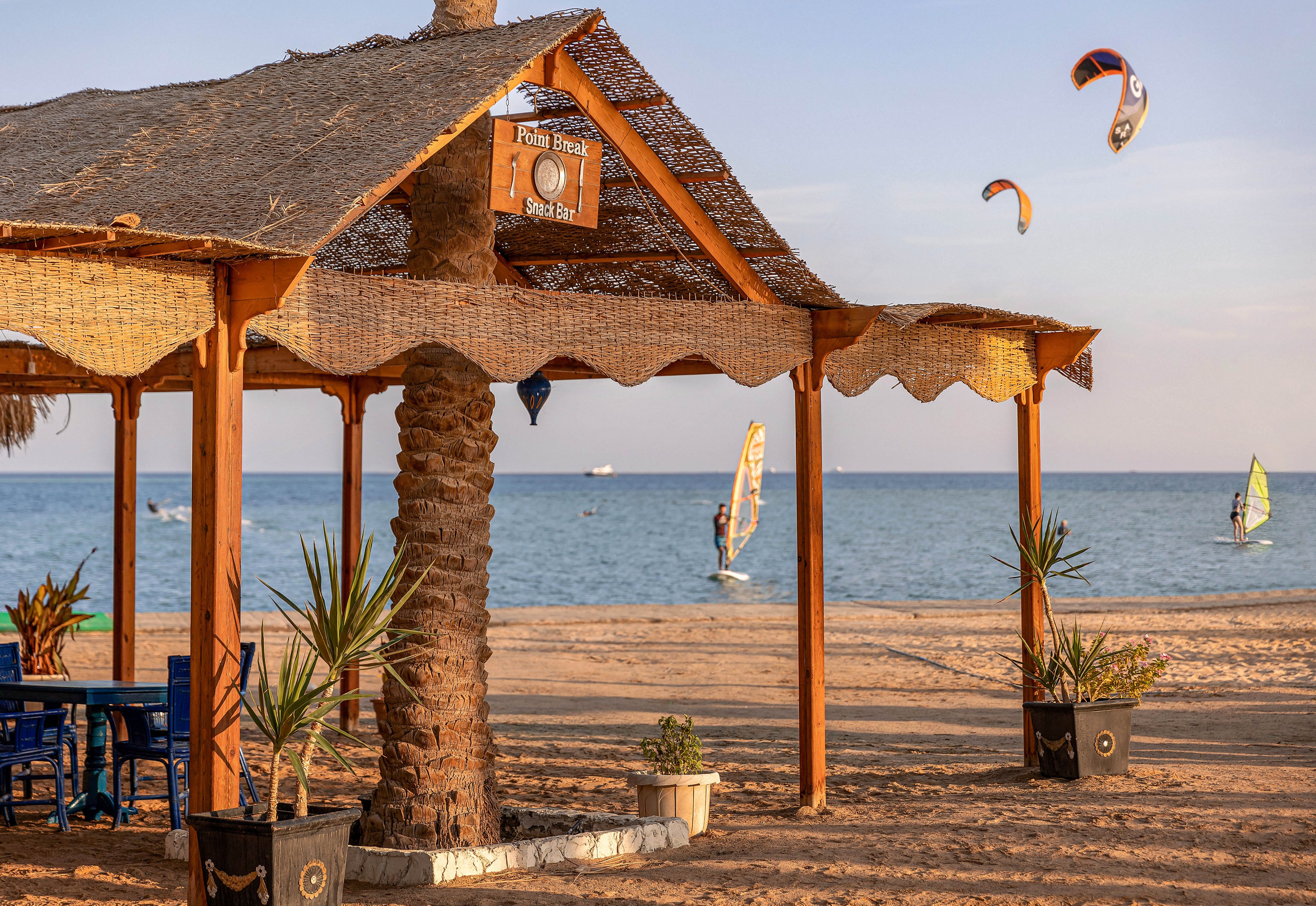 A rustic wooden hut stands on a sandy beach, shaded by a palm tree and surrounded by potted plants. In the background, several kite surfers glide across the blue sea under a clear sky. The scene is tranquil, with warm golden tones and a relaxed coastal atmosphere. A sign reading 'Family House Snack Bar' is visible above the hut entrance.