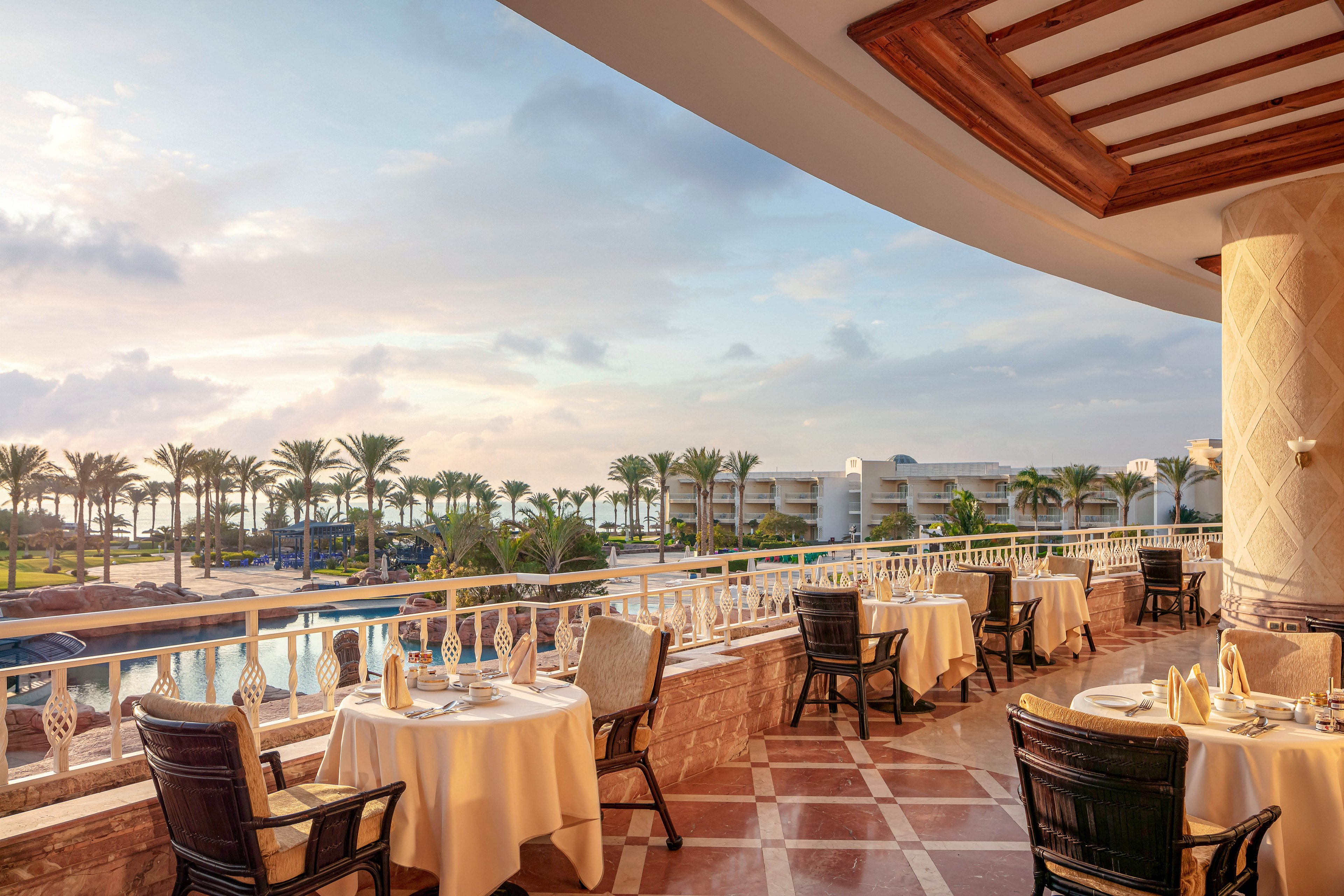 The image features a luxurious outdoor dining area set on a tiled terrace with neatly arranged tables and chairs. The setting overlooks a resort pool surrounded by palm trees, with hotel buildings visible in the background. The scene is bathed in warm, natural light, creating a serene and inviting atmosphere. No visible text or numeric values are present in the image.