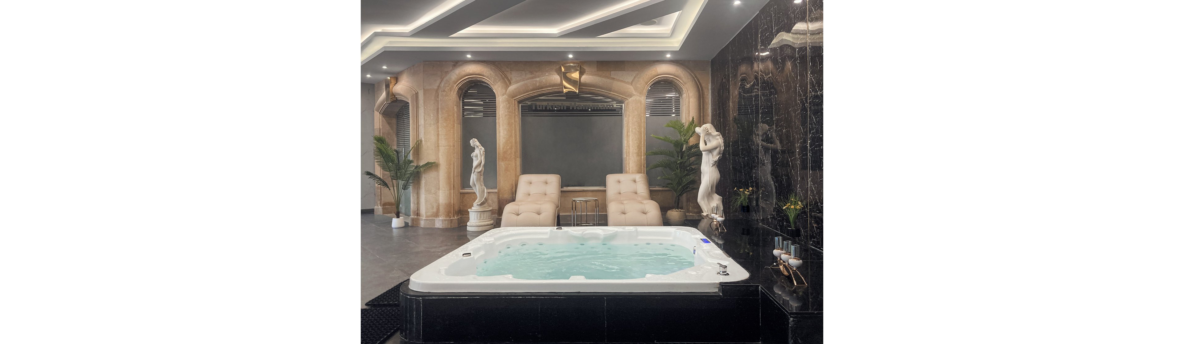 The image features a modern indoor spa setting with a bubbling jacuzzi at the center. Two elegant lounge chairs are positioned against a stone wall, flanked by classical statues and lush green plants. The space is illuminated by recessed ceiling lights, creating a serene and upscale atmosphere. No visible text or numeric values are present in the image.