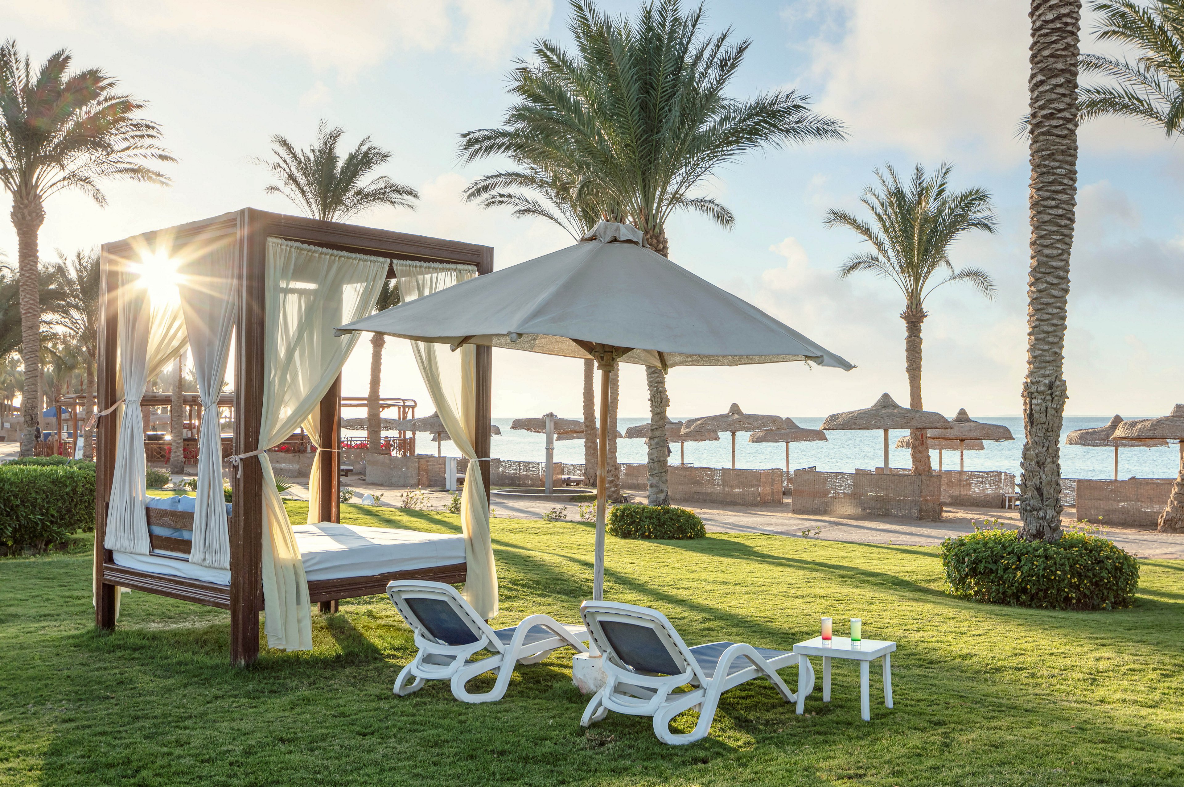 A stylish cabana with flowing curtains stands beside two white lounge chairs under a large umbrella on a grassy area. The scene overlooks a sandy beach with palm trees and straw parasols, with the sun rising in the background. Two drinks are placed on a small table, adding a touch of relaxation to the tranquil setting.