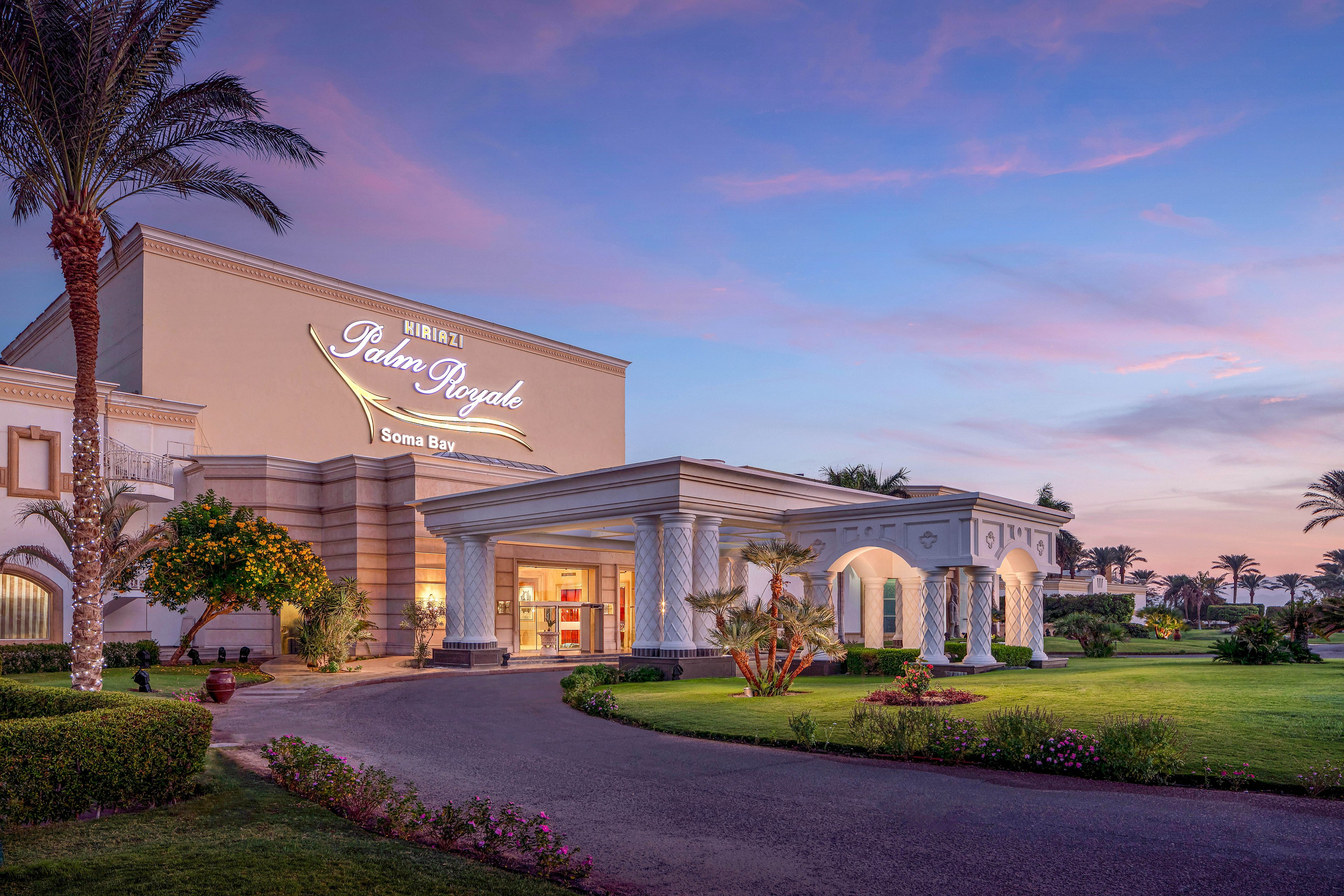 The image shows the grand entrance of the Kempinski Palm Royale hotel in Soma Bay, illuminated during sunset. The building features elegant columns and lush landscaping, with palm trees and manicured lawns enhancing the luxurious atmosphere. The sky is painted in soft hues of pink and purple, creating a tranquil and inviting mood. The hotel name and location are clearly visible on the facade.