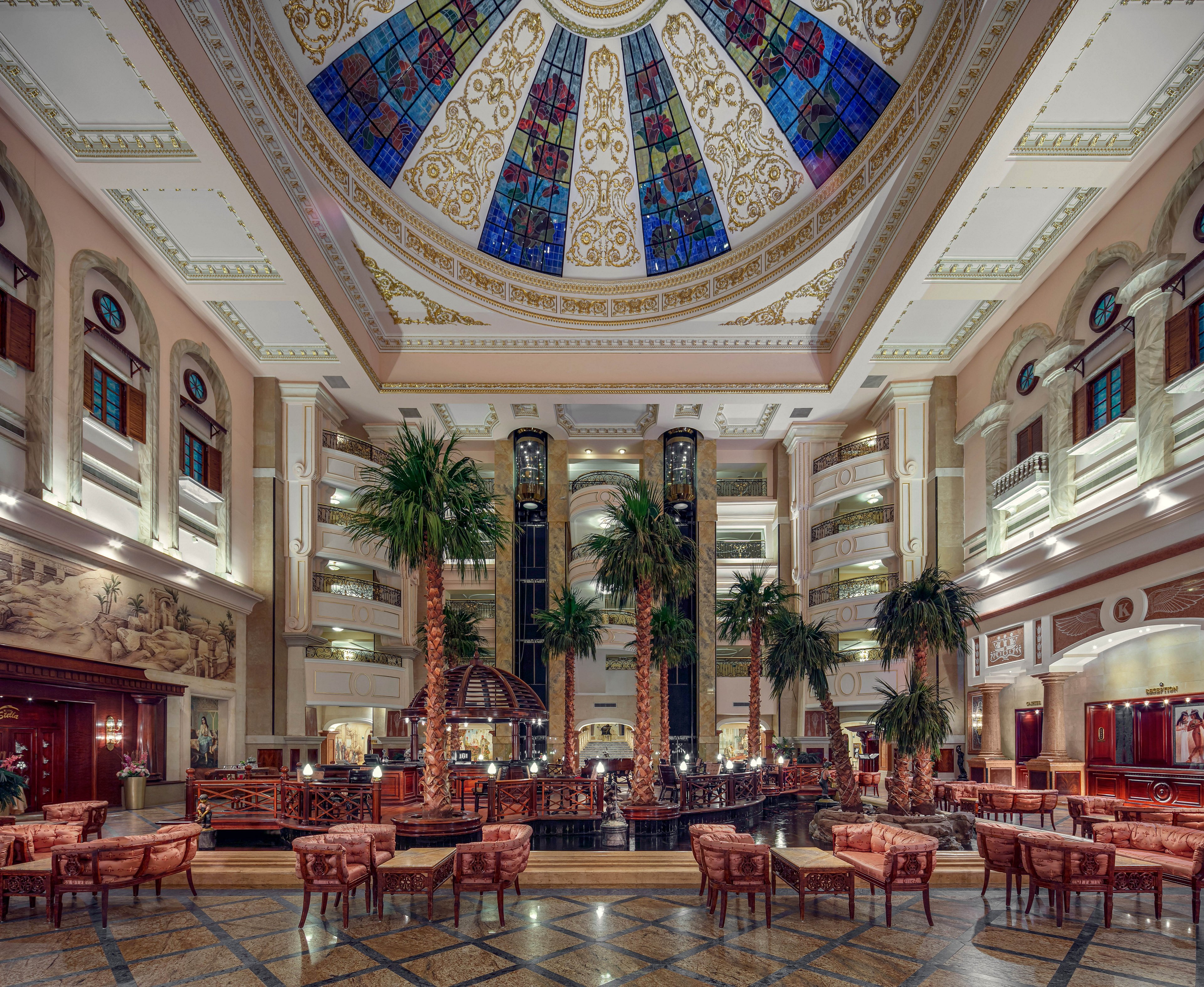 The image features an opulent hotel lobby with a grand stained glass dome ceiling as its centerpiece. Tall palm trees and elegant seating arrangements create a welcoming atmosphere, while marble floors and intricate architectural details enhance the sense of luxury. The space is brightly lit, with warm tones and classical decor contributing to a sophisticated mood. No visible text or numeric values are present in the image.