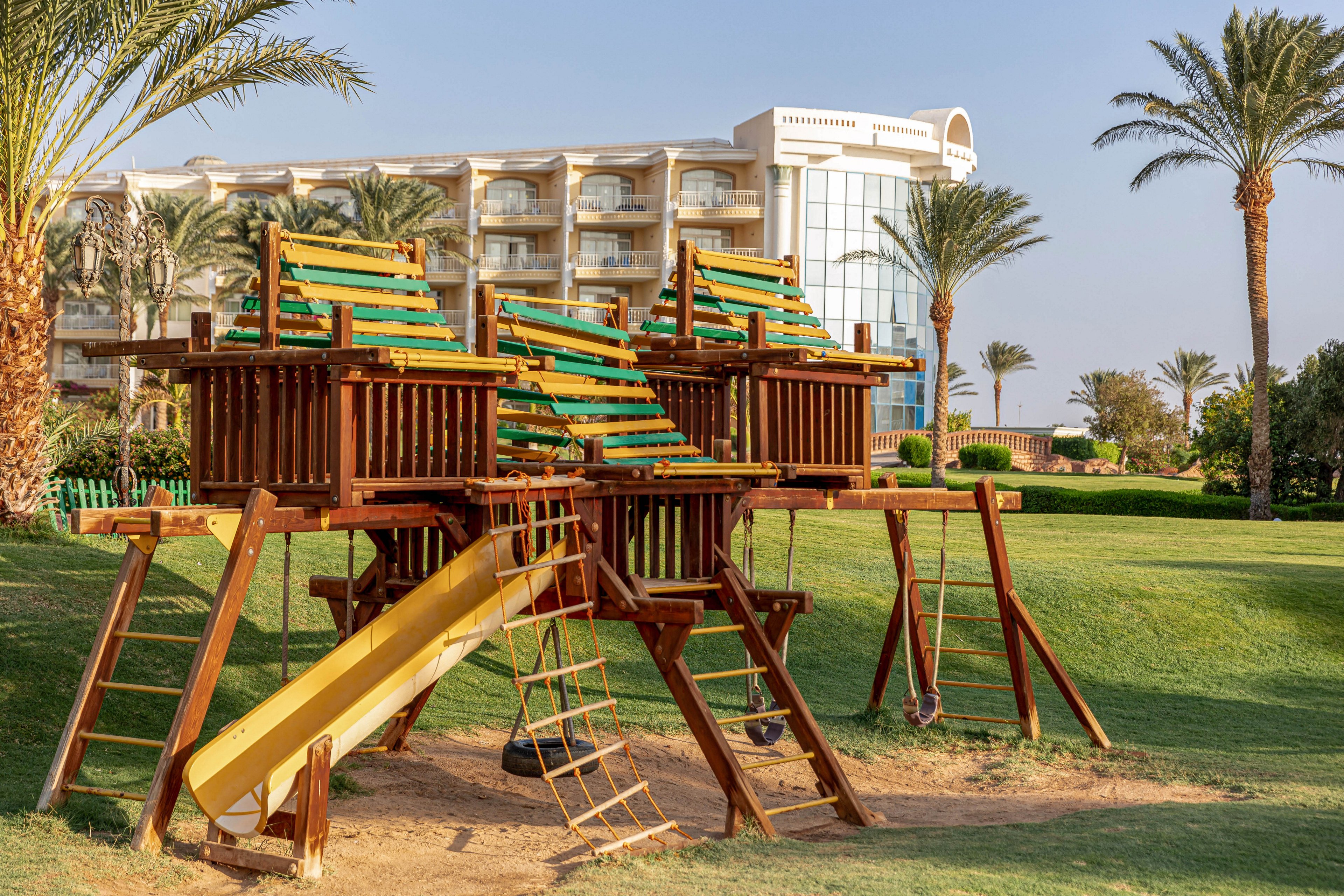 A wooden playground with slides, ladders, and climbing frames stands on a grassy area in front of a modern hotel building. The scene is set outdoors with palm trees and clear skies, suggesting a warm, sunny day. The playground features yellow and green accents, and the hotel architecture is visible in the background. No visible text or numbers are present in the image.