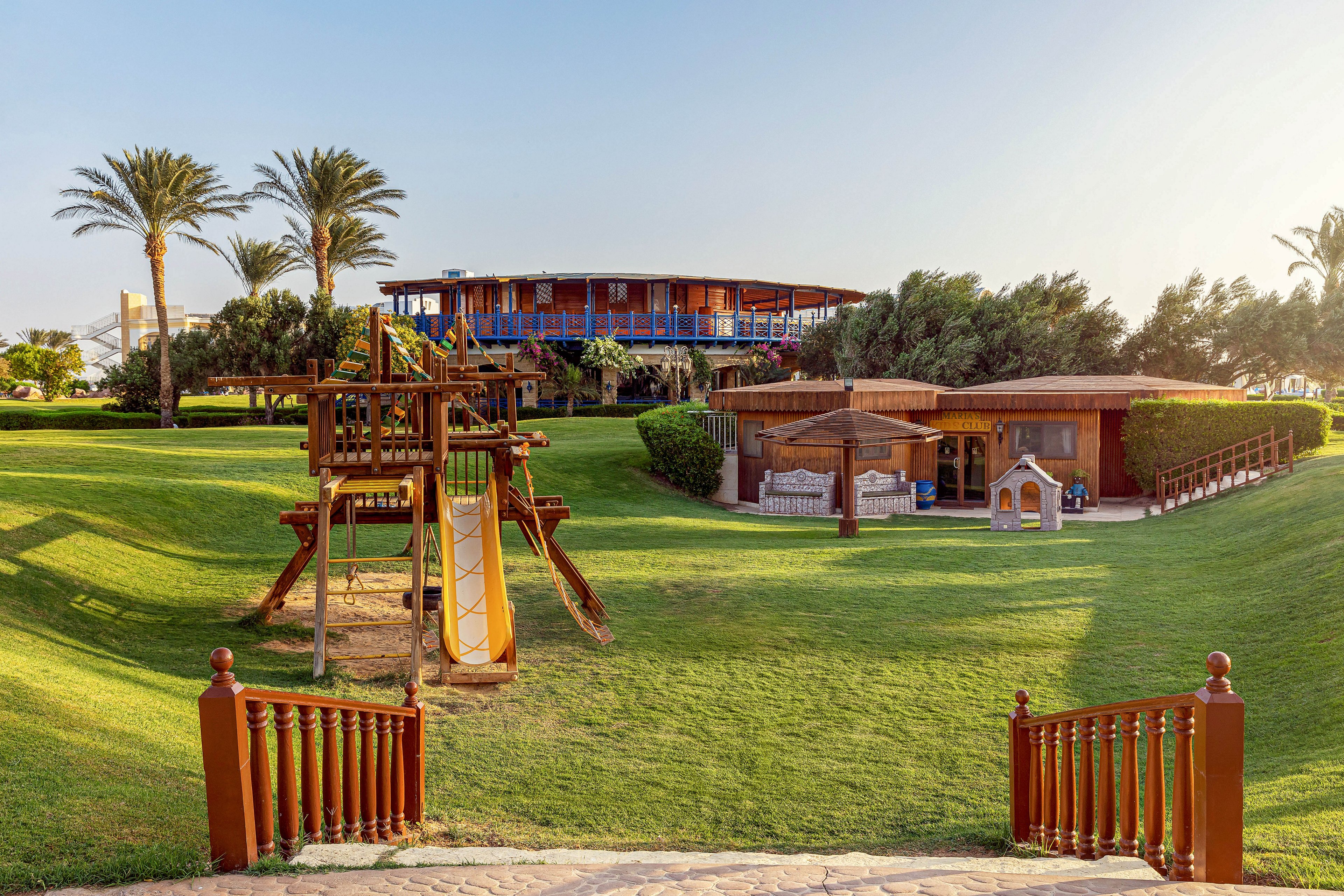A well-maintained playground features a wooden climbing frame with a yellow slide, set on lush green grass. The area is surrounded by palm trees and wooden buildings, suggesting a resort or recreational park setting. The lighting is warm and natural, creating a cheerful and inviting mood. No visible text or numeric values are present in the image.