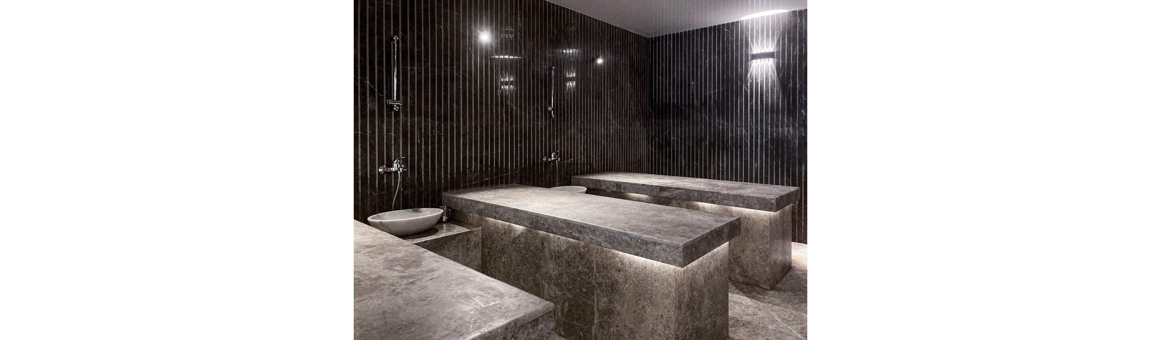 A contemporary spa room features large stone massage tables illuminated from below, creating a dramatic effect. The walls are covered in dark tiles with vertical lines, and the space includes a modern sink and wall-mounted shower fixtures. The lighting is soft and moody, enhancing the luxurious and tranquil atmosphere. No visible text or numbers are present in the image.